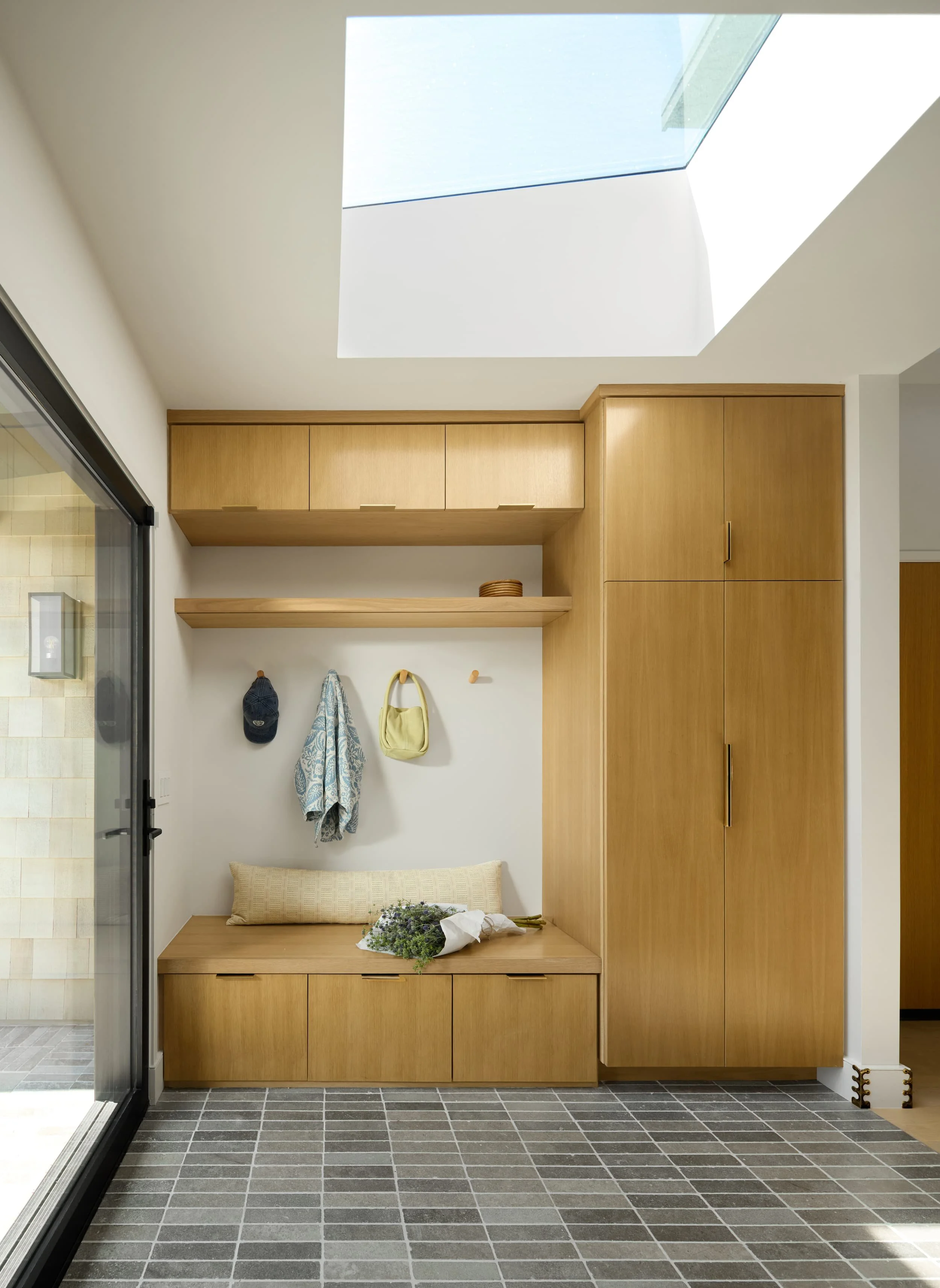 Mudroom with white oak built-in bench and cabinets under a skylight, with gray tile floor
