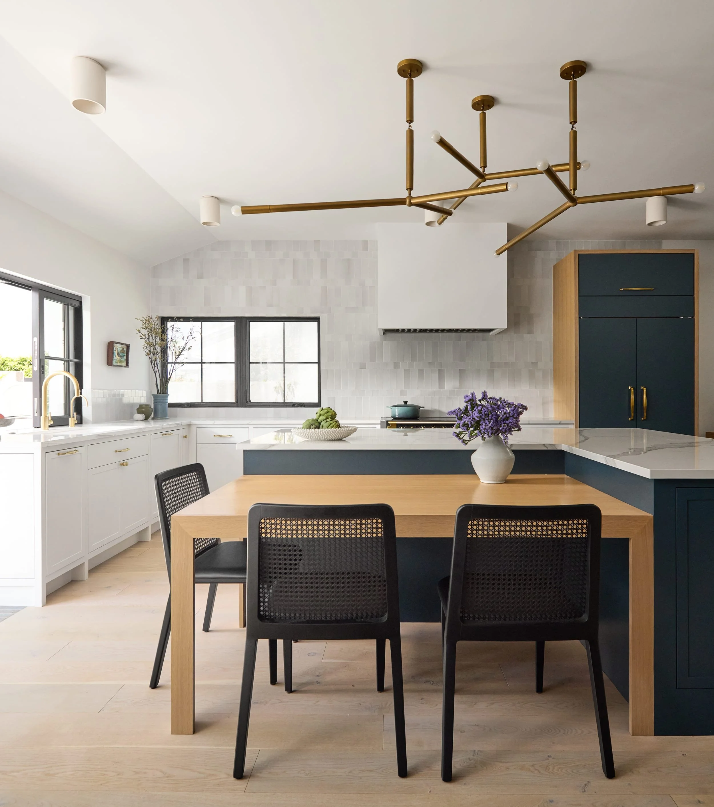 Kitchen with a modern brass chandelier over an oak dining table, navy island and white plaster range hood
