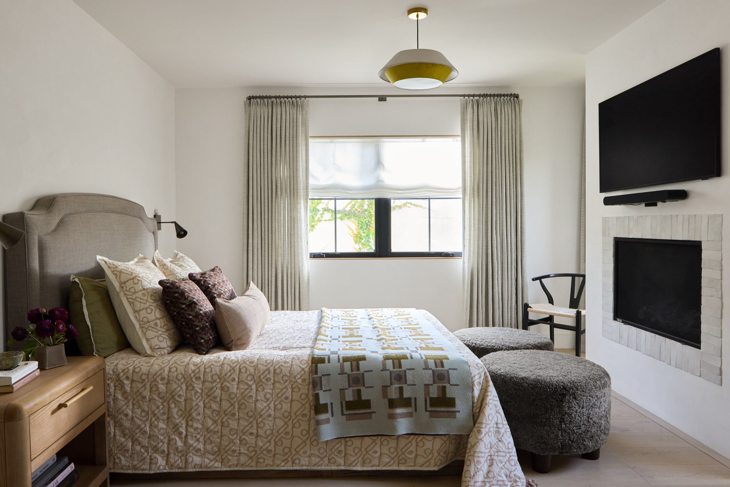 Bedroom with an upholstered headboard, layered neutral bedding, linen drapes and a tiled fireplace