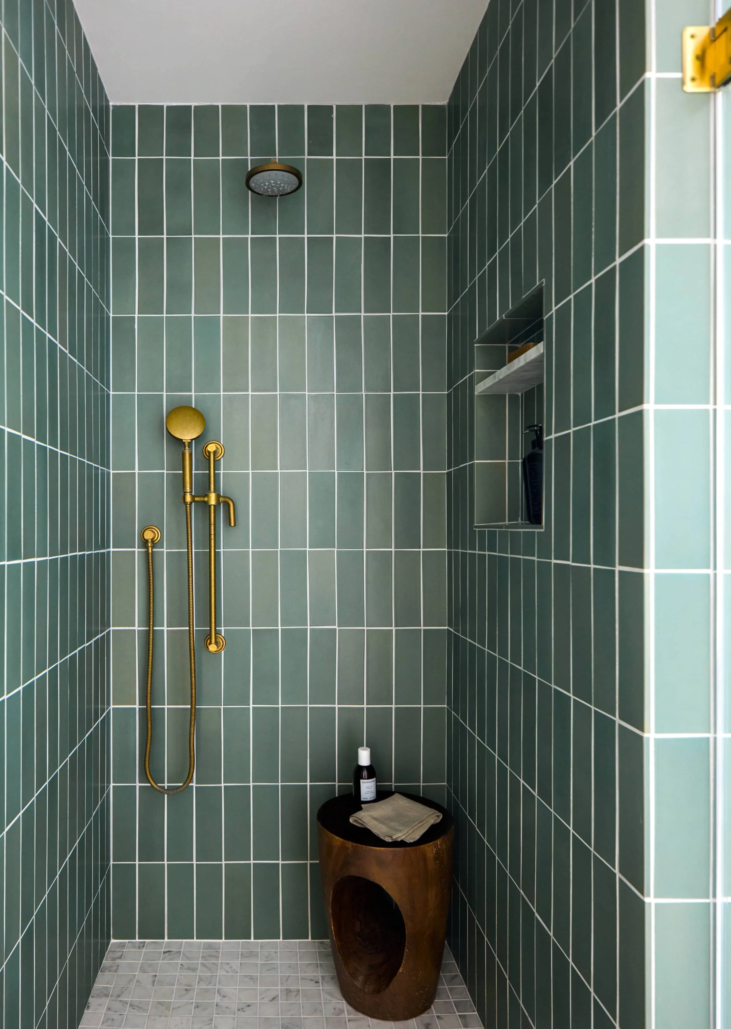Walk-in shower with green vertical tile, brass fixtures and a carved wood stool