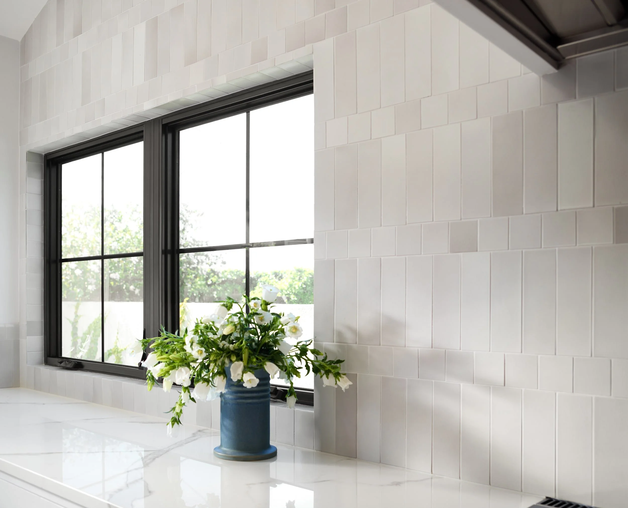 Black-framed kitchen window with stacked heath tile, marble counter and a blue vase of white flowers