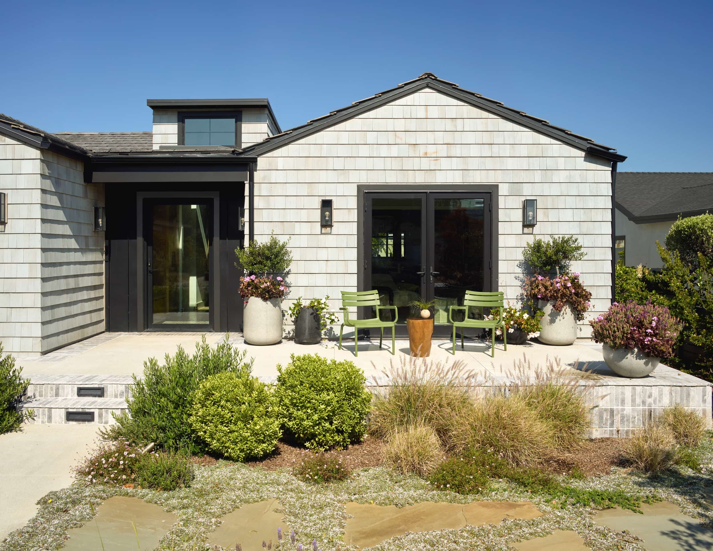 Modern shingle home exterior with black doors, potted plants and a patio with green chairs