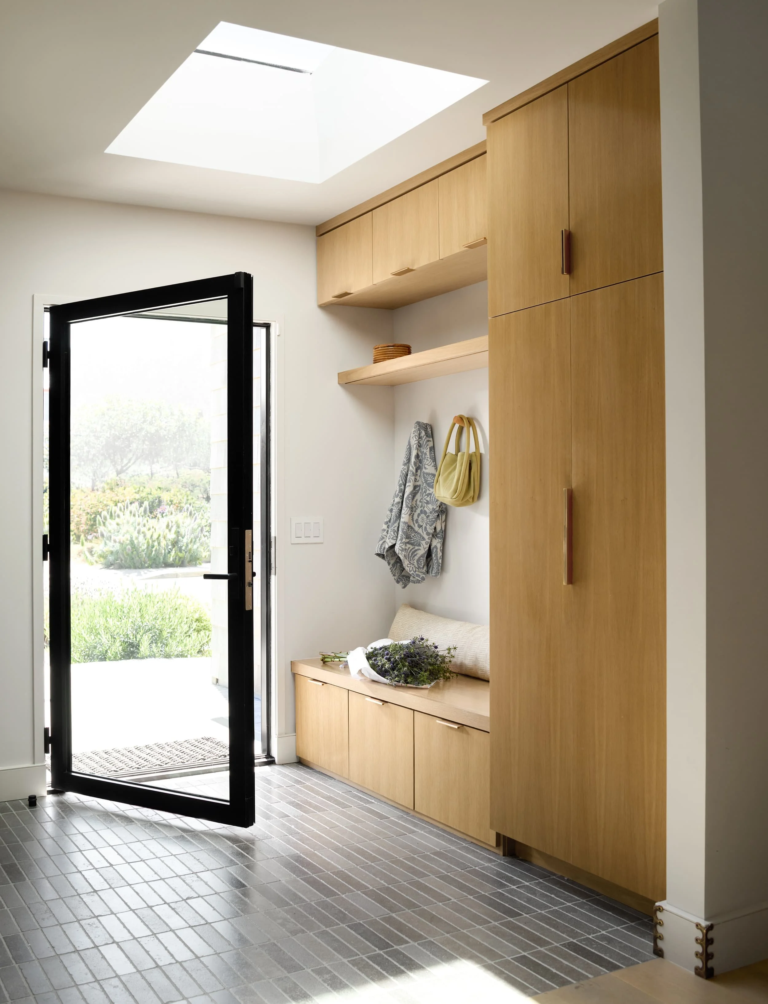 Entry with a black steel glass door opening to a white oak mudroom bench and storage under a skylight