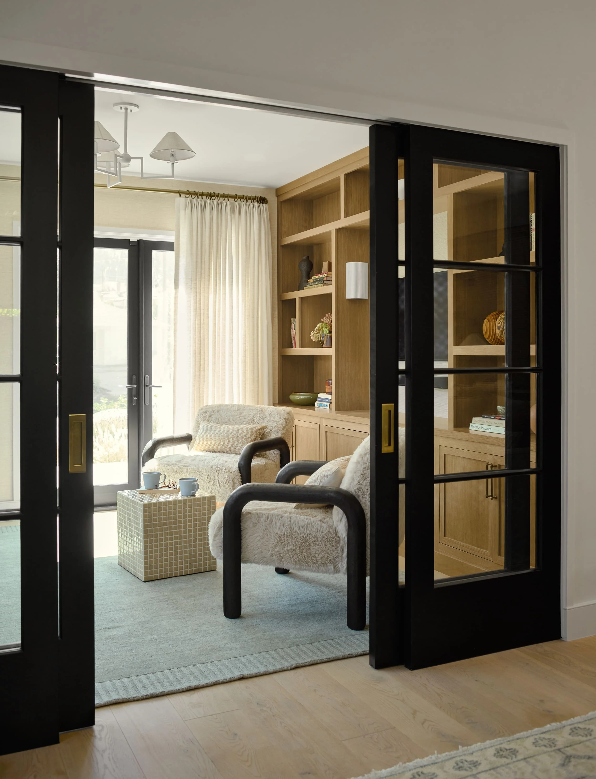 Black glass French doors opening into a den with shearling chairs and white oak built-ins