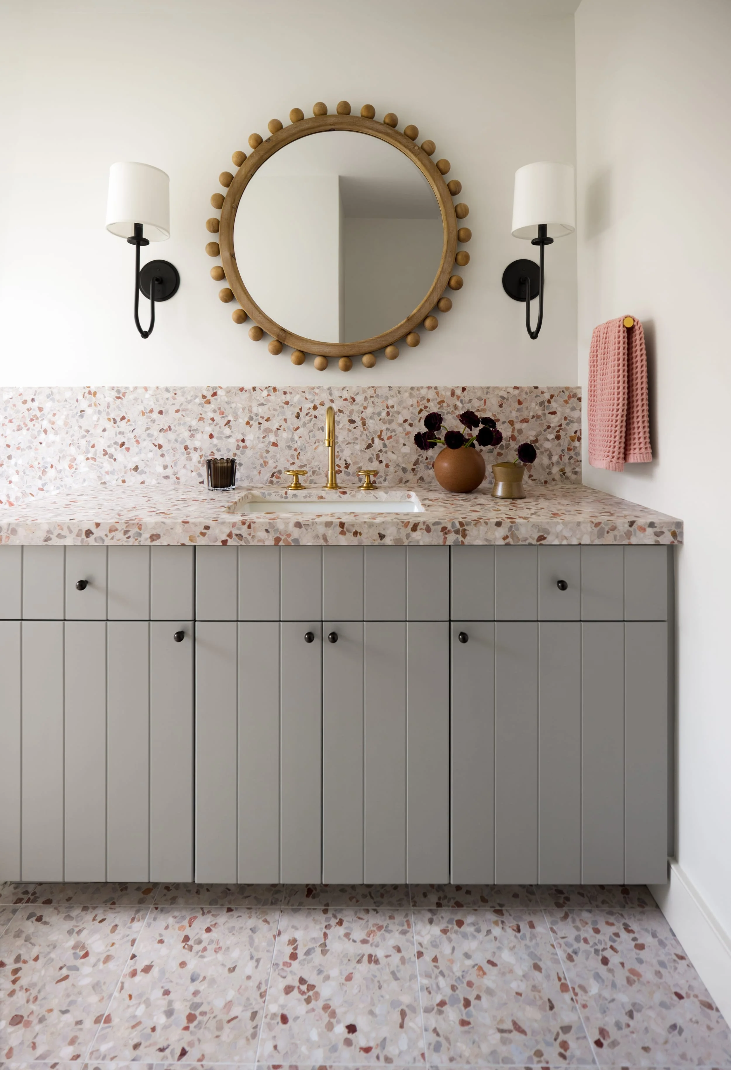 Bathroom with a pink terrazzo backsplash and floor, gray vanity and round beaded brass mirror
