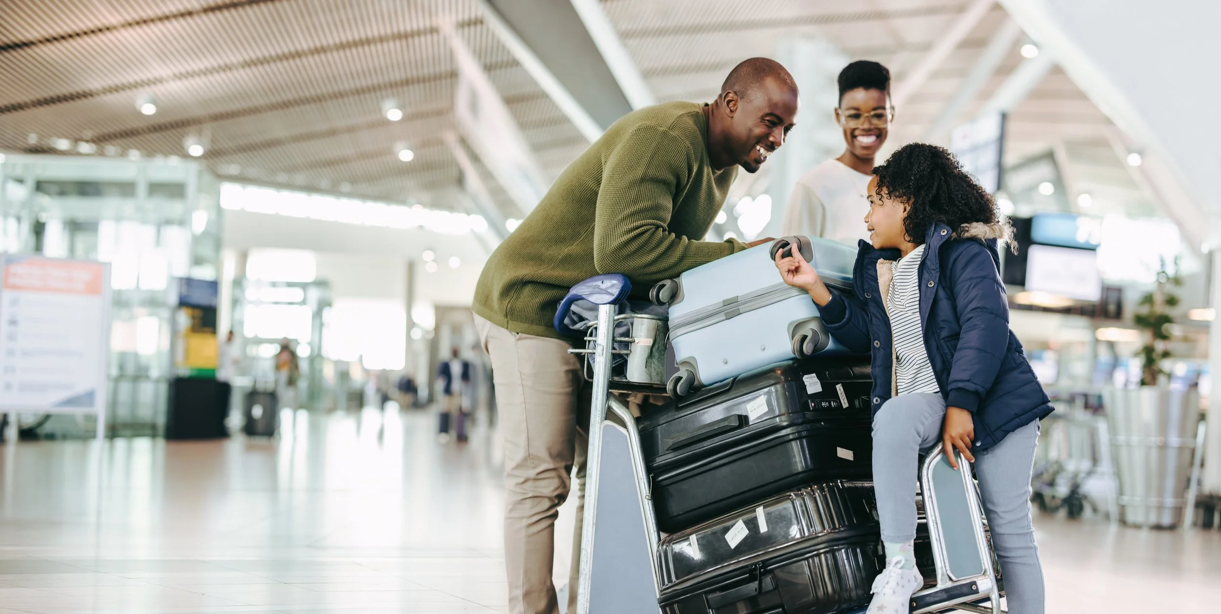 A family of three at an airport, engaged in a conversation with a cart holding multiple suitcases. The father is leaning over the cart, smiling, while the young daughter in a blue jacket talks to him. The mother is standing behind, smiling.