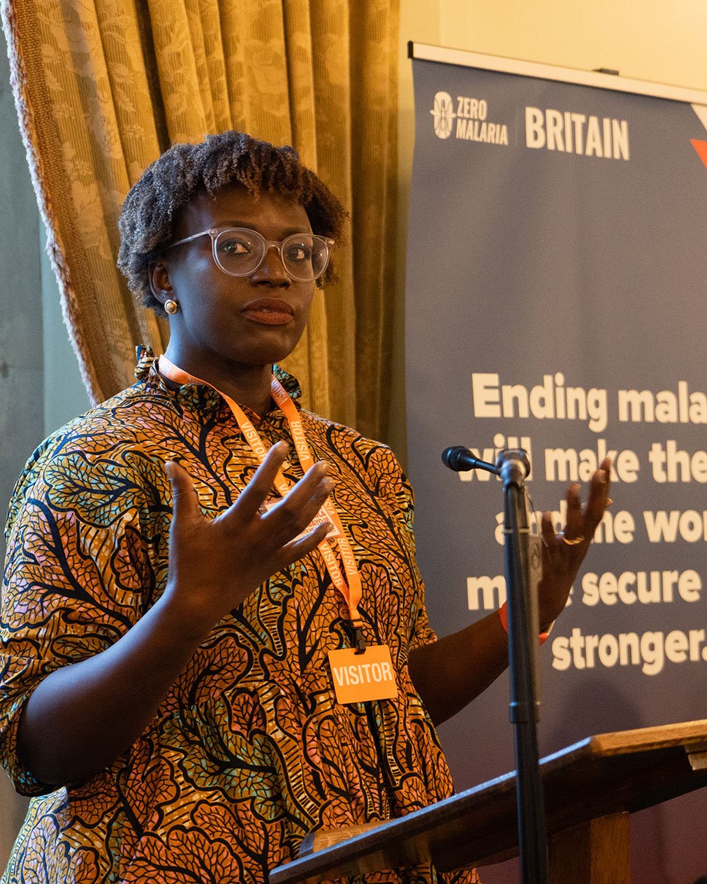 A woman with short, curly hair wearing glasses and a brightly patterned orange and yellow dress, speaking at a podium with a microphone during an event about malaria in Britain, with a banner in the background highlighting the organization's goal to end malaria.
