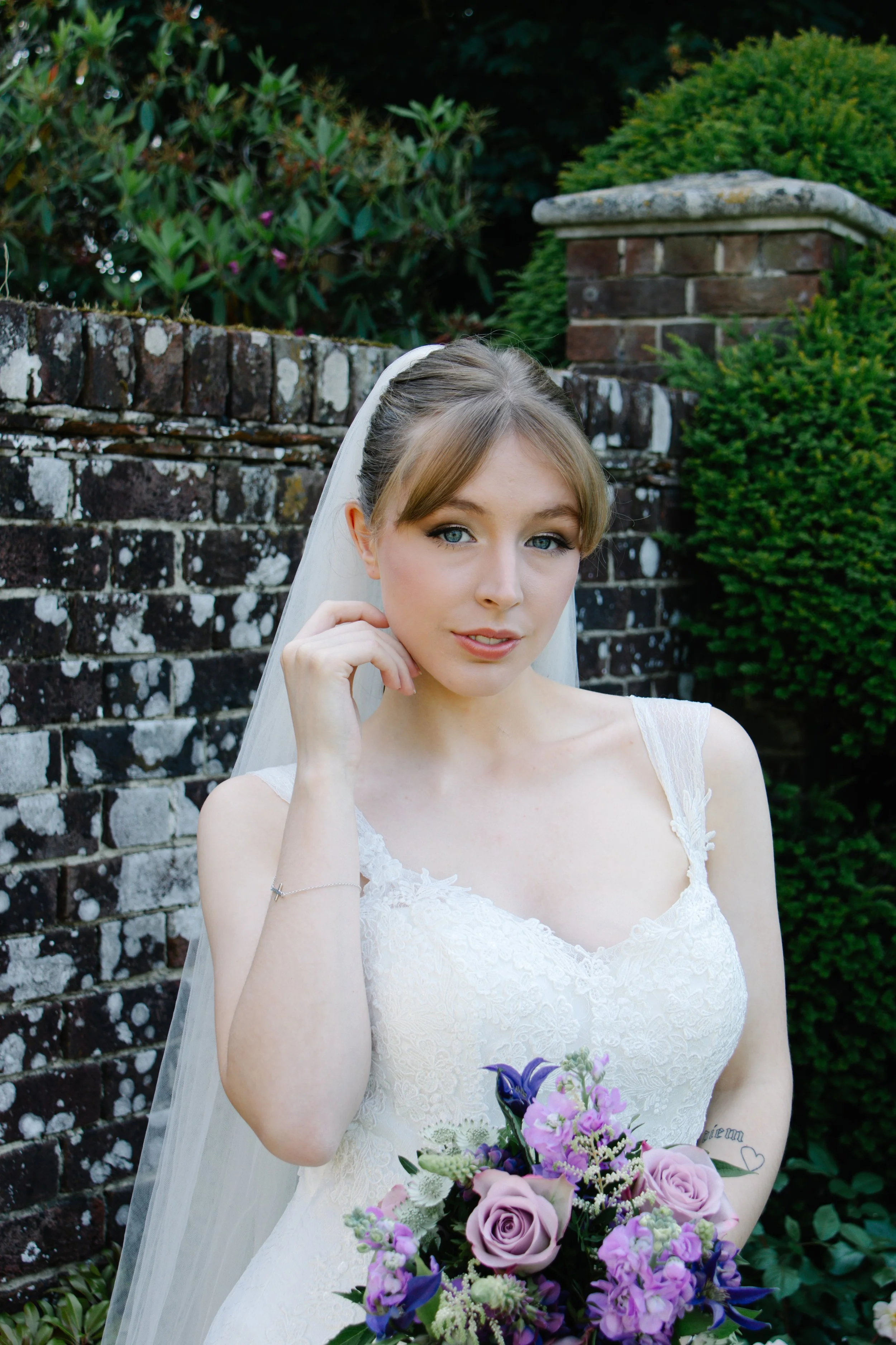 A bride in a white lace wedding dress holding a bouquet of purple and pink flowers, standing outdoors near a brick wall and green bushes.