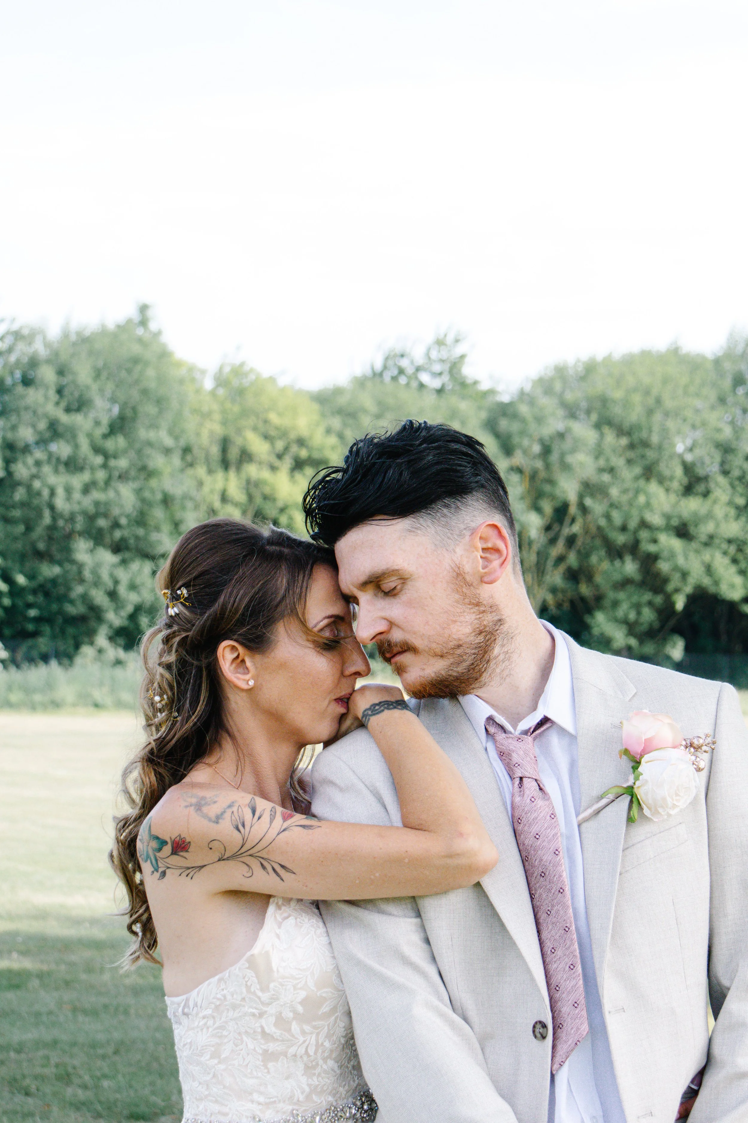 A bride and groom in a close embrace during their wedding, outdoors with trees in the background. The bride has tattoos on her arm, and the groom is wearing a light gray suit with a pink tie and a pink boutonniere.