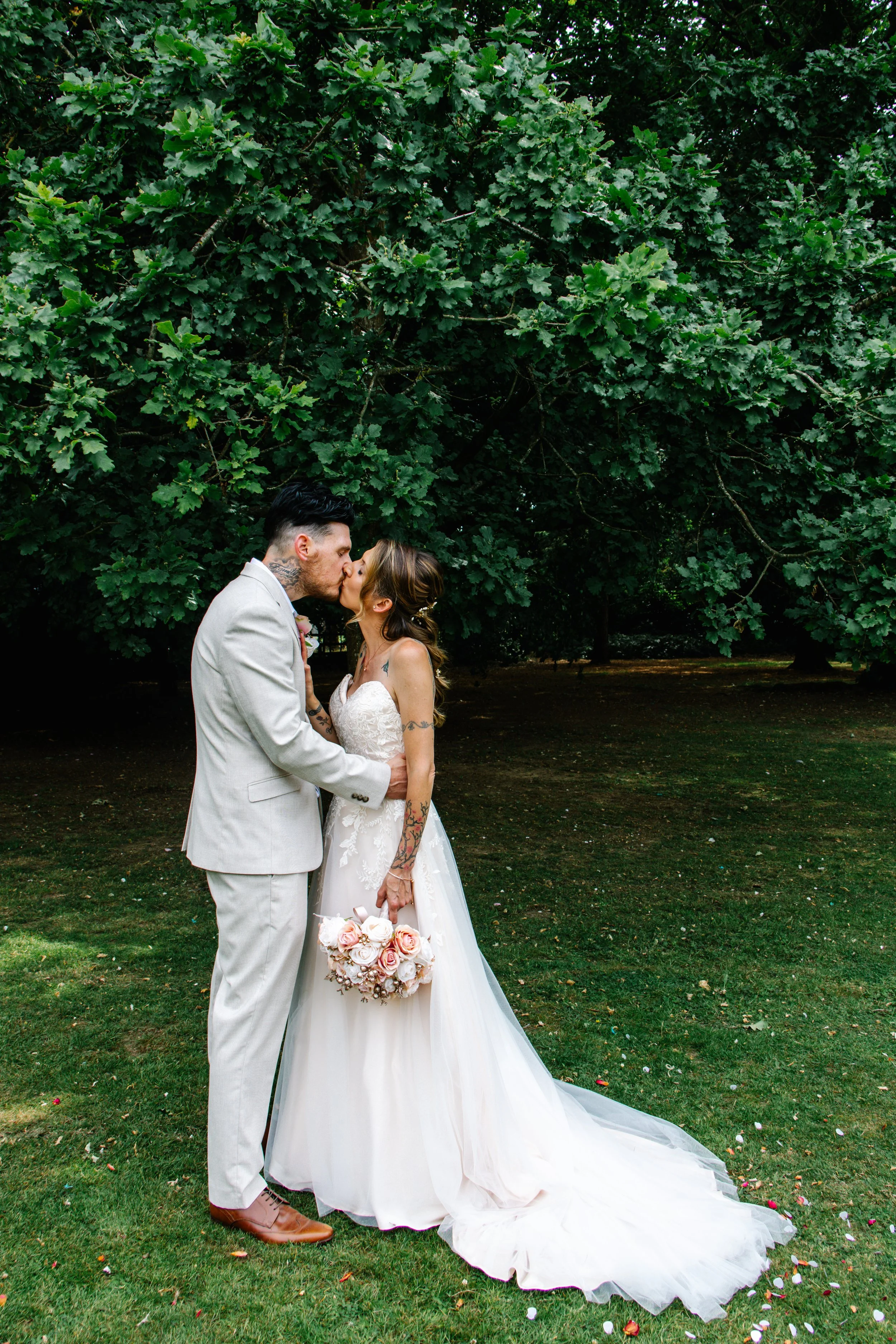 A bride and groom kiss outdoors under a large green tree, with the bride holding a bouquet of pink and white roses.