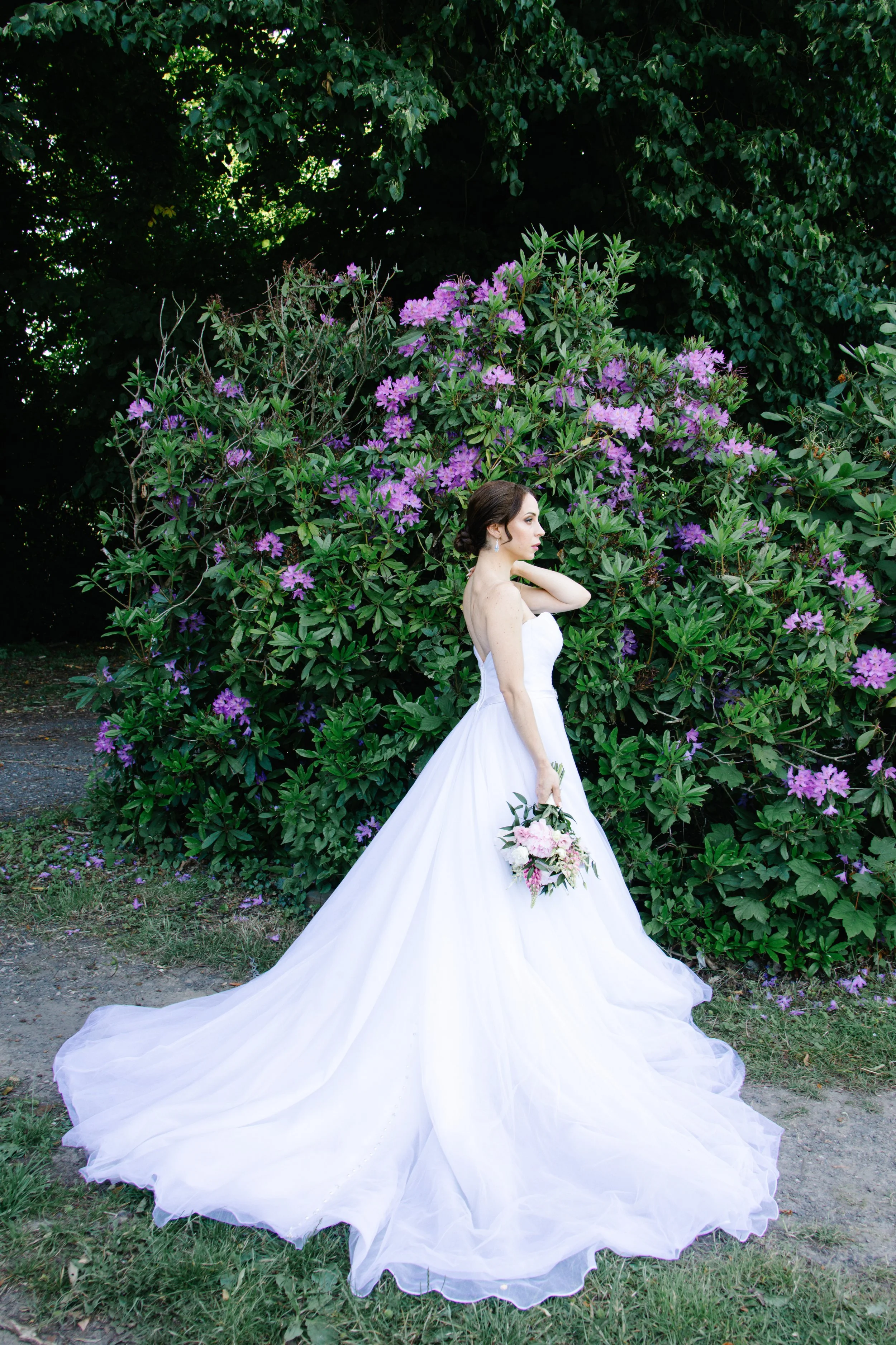 A bride in a white wedding gown holding a bouquet of pink and white flowers, standing outdoors in front of purple blooming bushes.