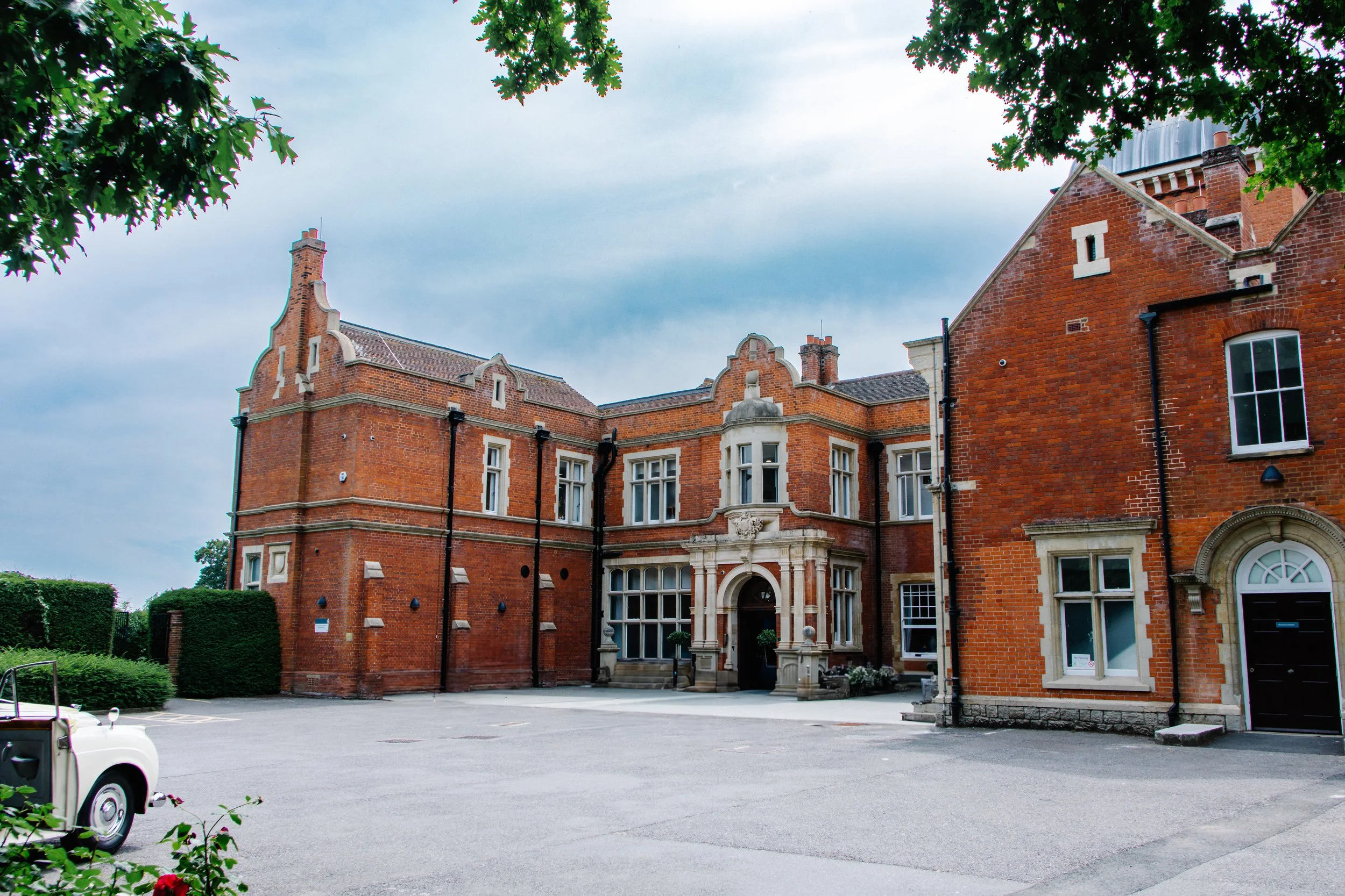 Large red brick building with ornate architecture, multiple windows, and a curved entrance, surrounded by a parking lot and greenery under a cloudy sky.