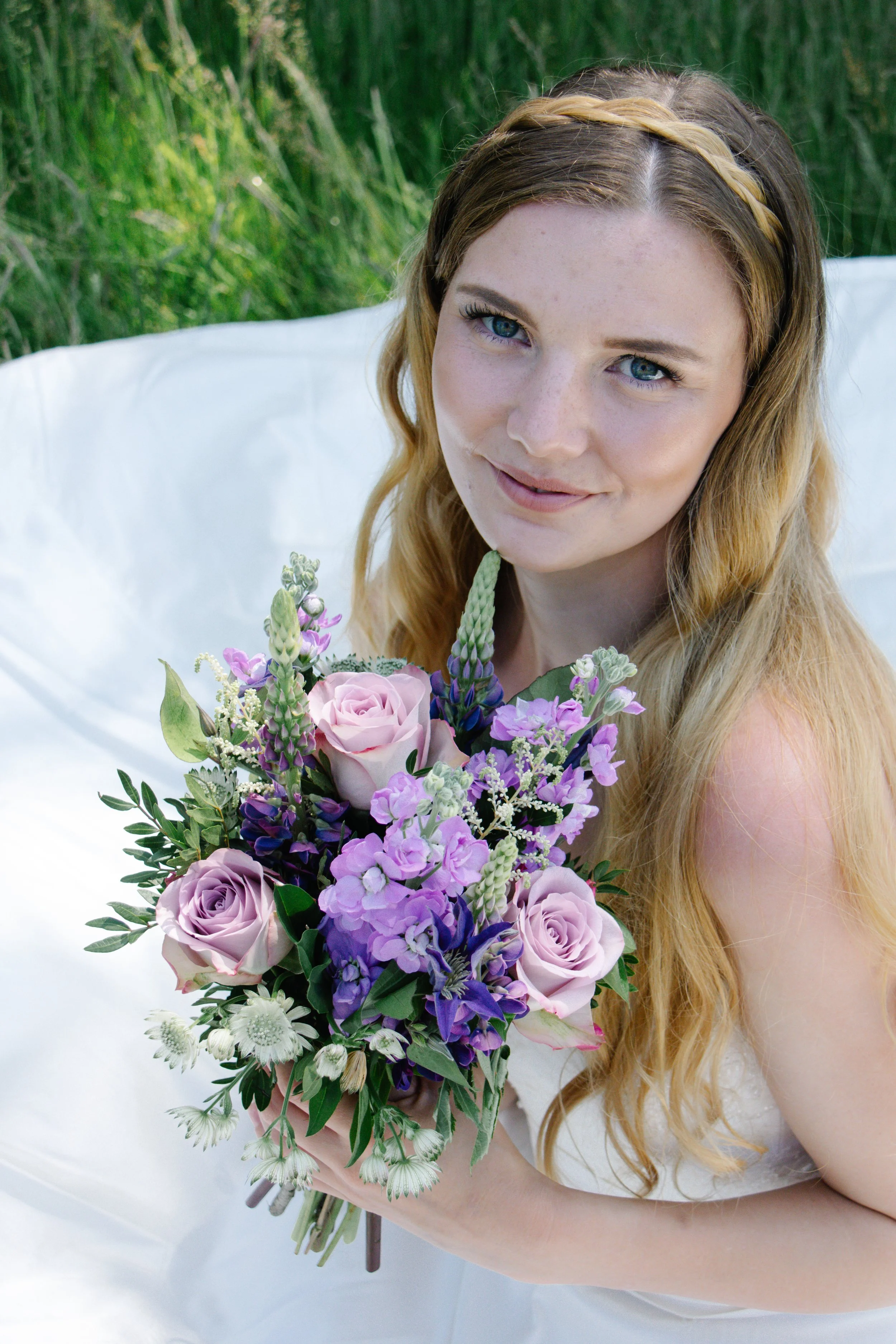 Young woman with long, wavy blonde hair, wearing a white dress, holding a bouquet of pink, purple, and white flowers, sitting outdoors on a white blanket, with green grass in the background.