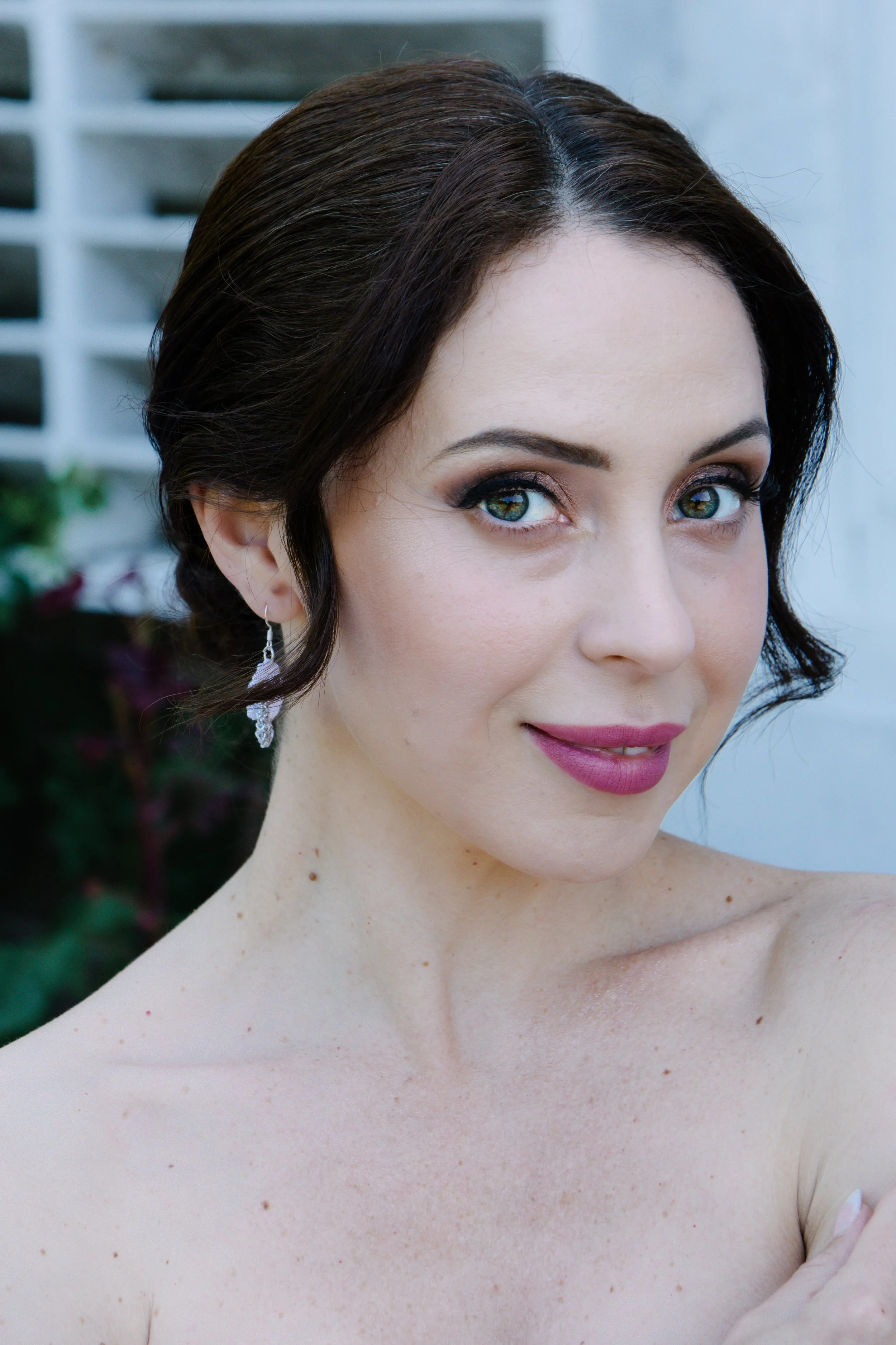Close-up of a woman with fair skin, dark brown hair styled in waves, wearing makeup with dark eyeshadow and pink lipstick, and dangling earrings, outdoors with a blurred background.
