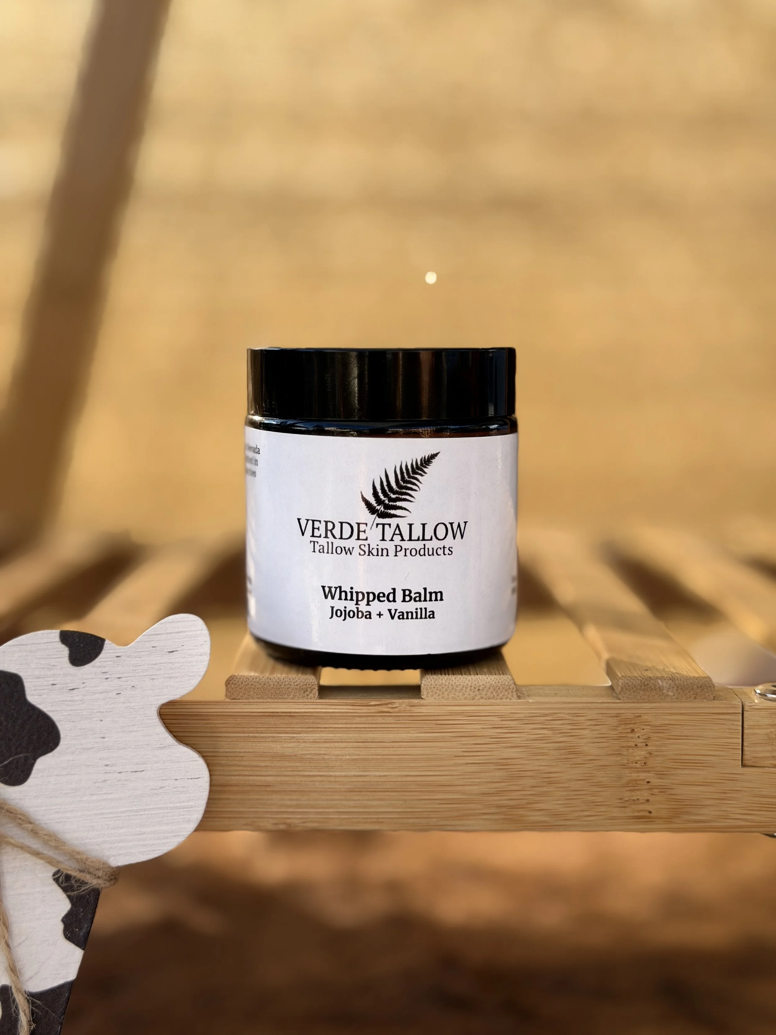 A jar of Verde Tallow whipped balm with jojoba and vanilla sitting on a wooden surface with a blurred background.
