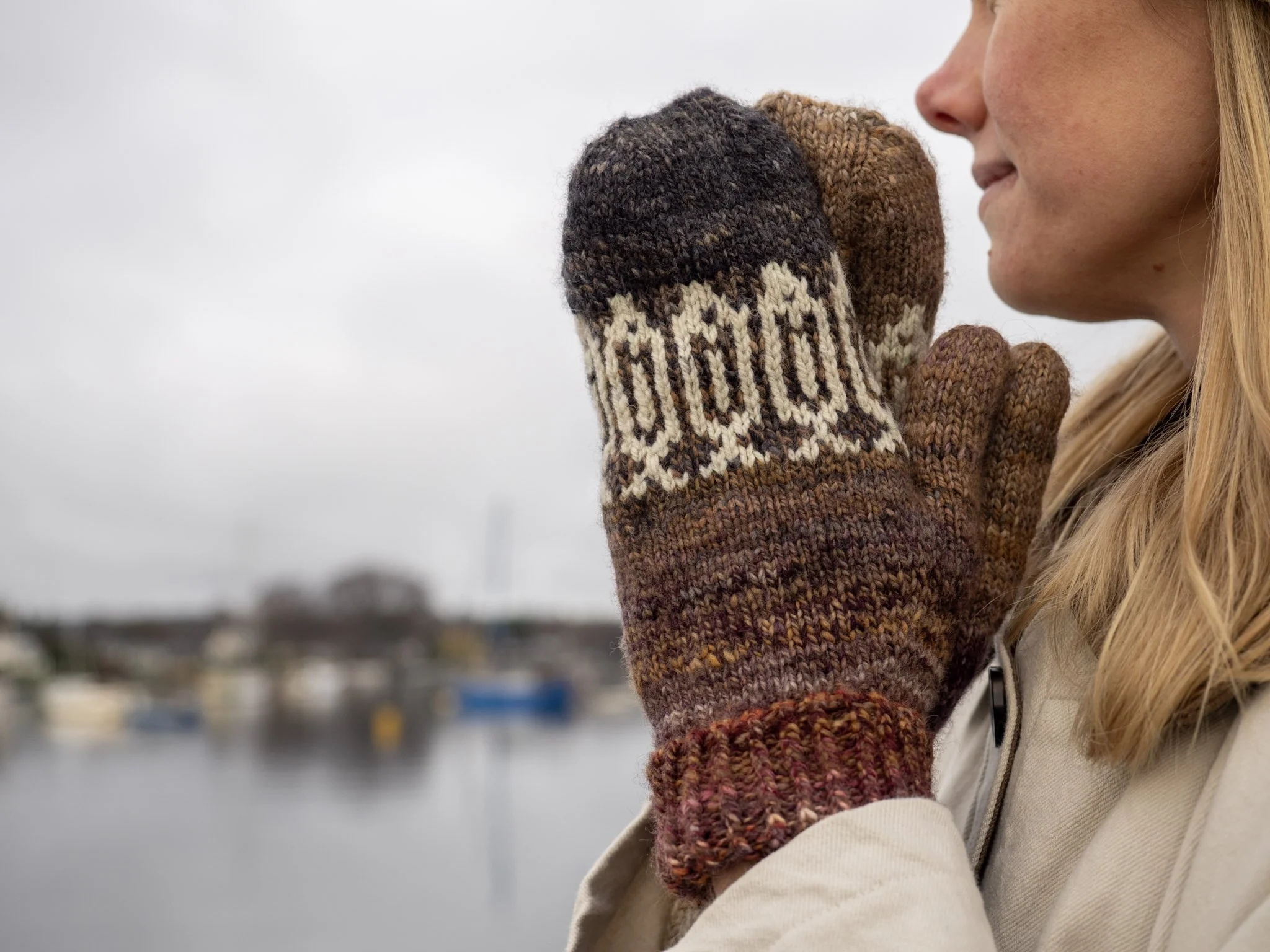 A woman wearing knitted mittens with a sardine fish pattern, holding her hands close to her face outdoors near a body of water, with a cityscape and overcast sky in the background.