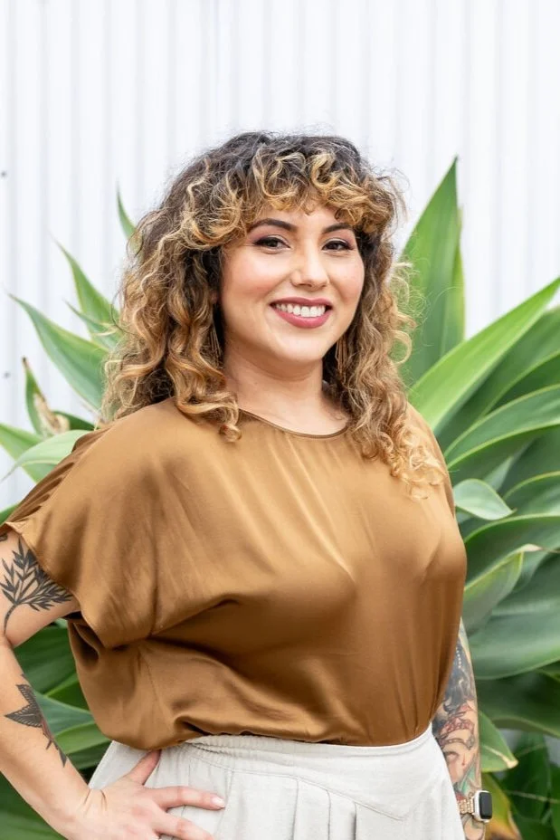 A smiling woman with curly hair and tattoos on her arms, wearing a brown satin top and light-colored pants, standing outdoors in front of large green plants.