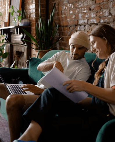 Two people sitting on a teal couch in a cozy room, looking at a notebook together, with a laptop nearby.