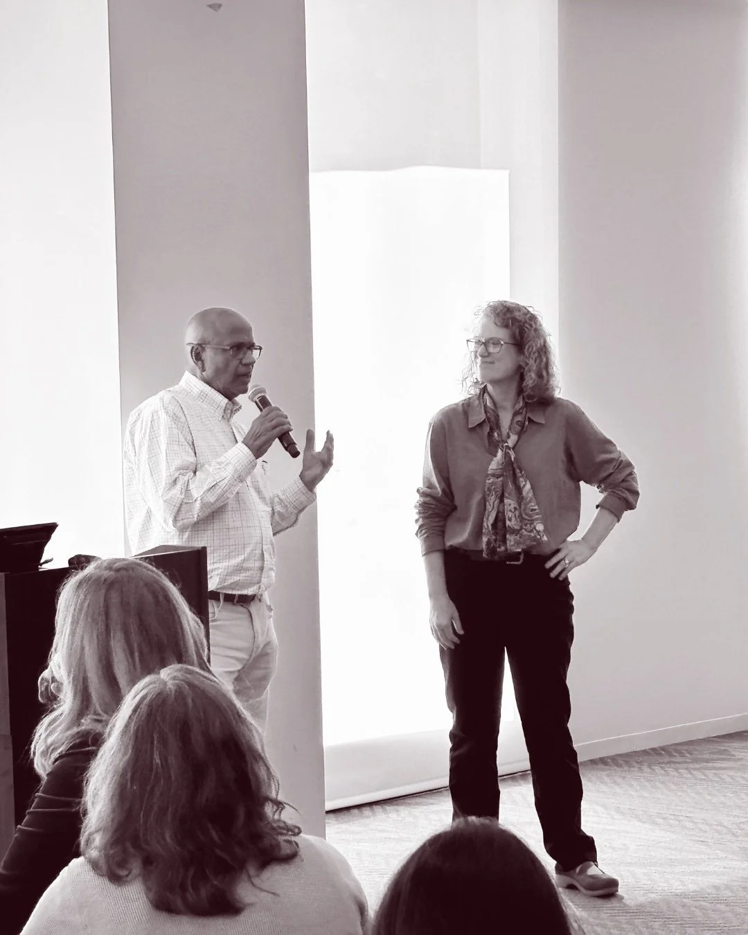 A man with glasses and a woman with curly hair speaking and listening during a presentation or discussion in a bright room with an audience.