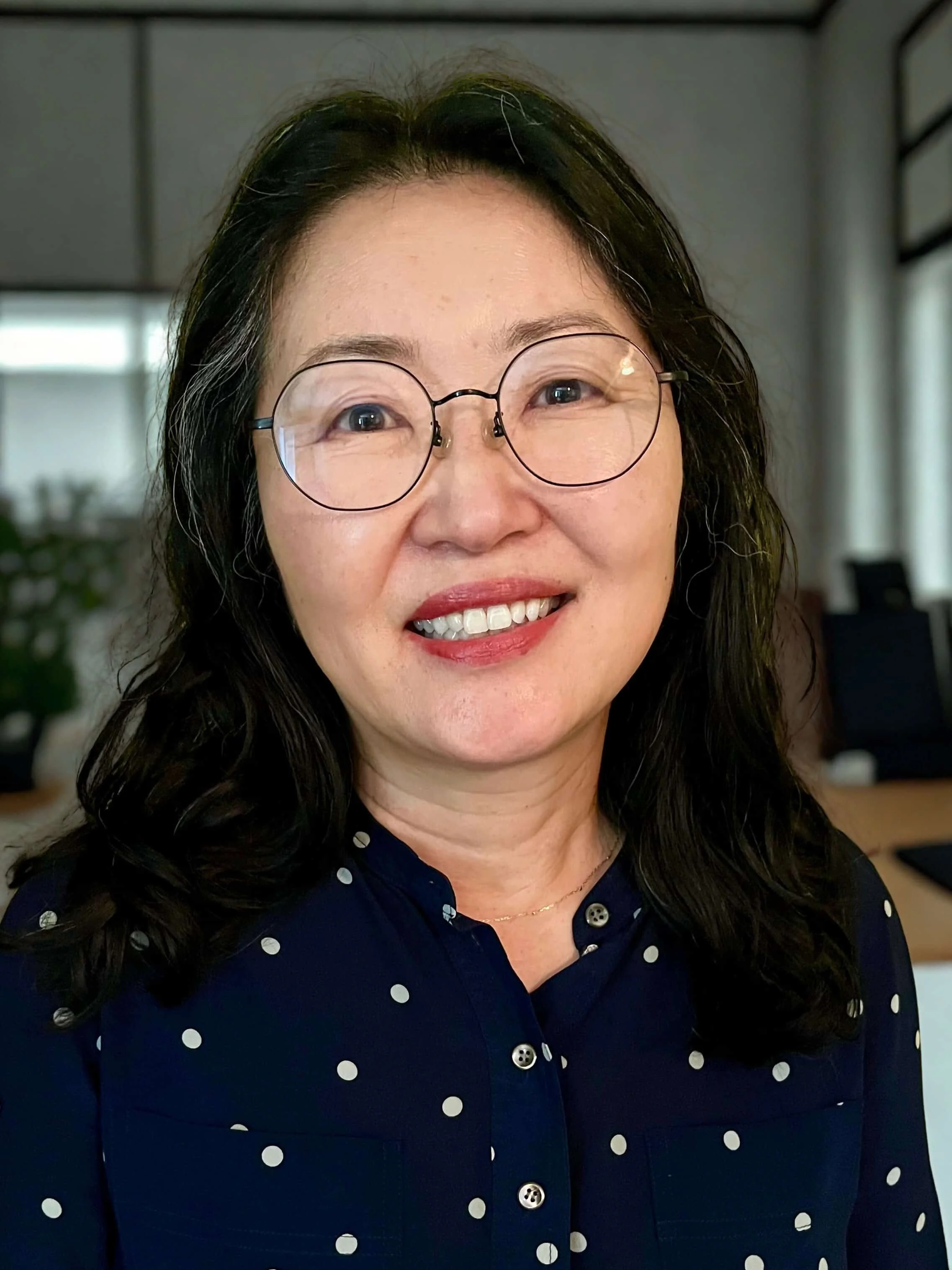 Close-up of an Asian woman with glasses, smiling, wearing a dark blue polka-dot shirt, in an office setting.