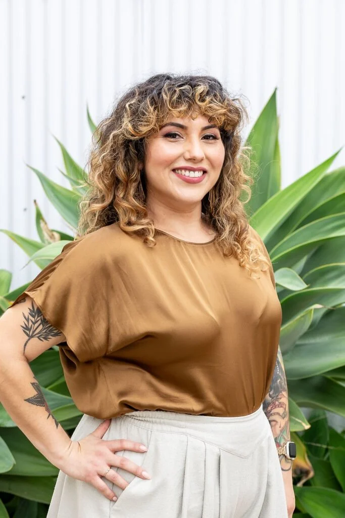 A woman with curly, shoulder-length hair and tattoos on her arms, smiling and standing outdoors in front of large green plants, wearing a brown silky top and light-colored pants.