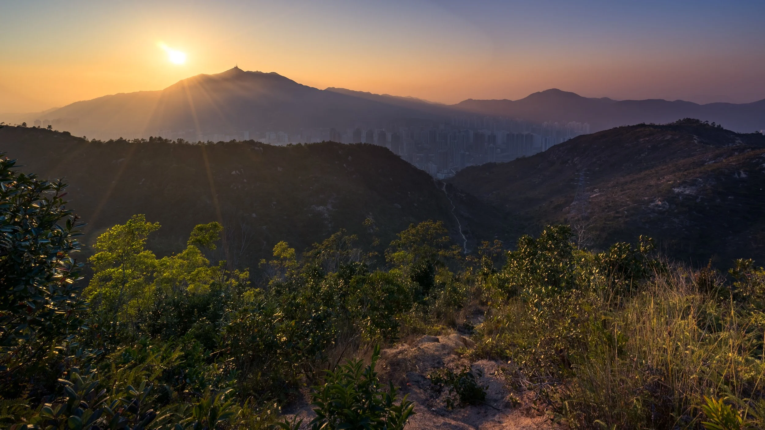 Sunset over mountainous landscape with city buildings visible in the distance, lush vegetation in foreground, and a lightning strike in a valley.