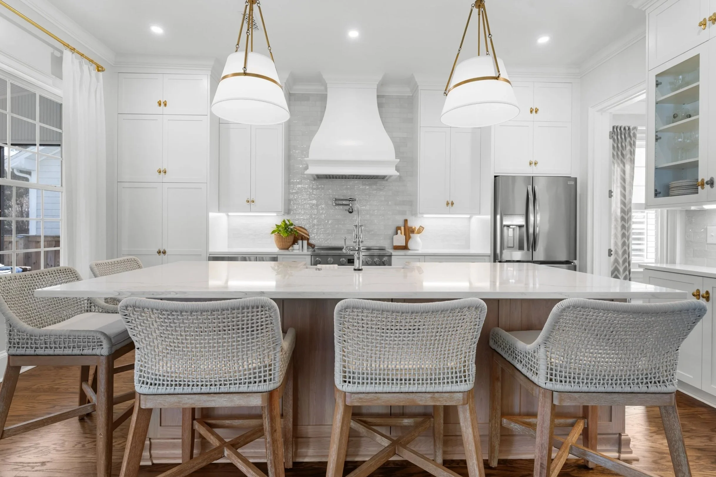Bright, white kitchen with a large island and woven bar stools, white cabinetry, stainless steel refrigerator, and a window with white curtains.