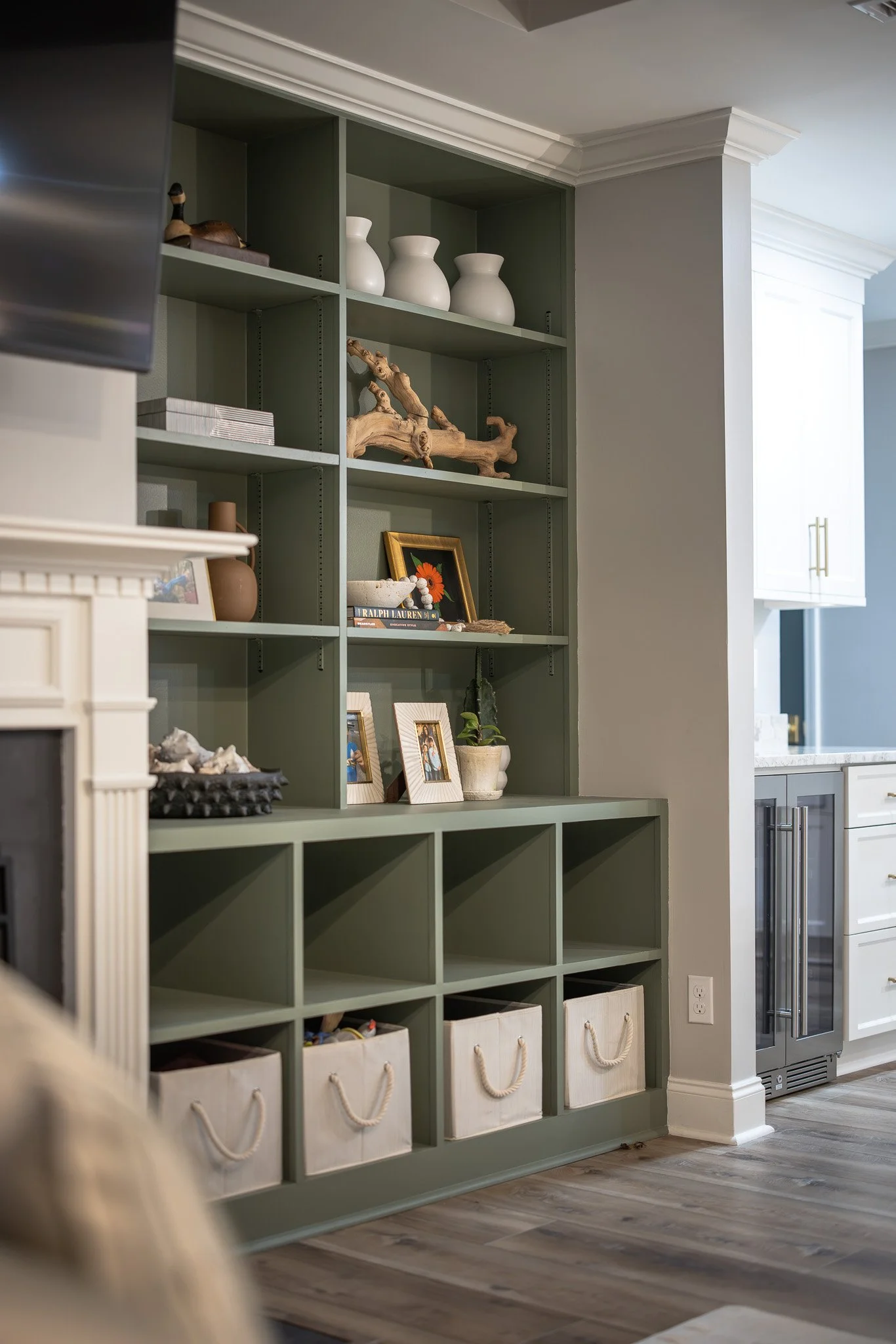 Green built-in bookshelf with decorative items, framed photos, and white storage bins, against a light gray wall in a modern kitchen.