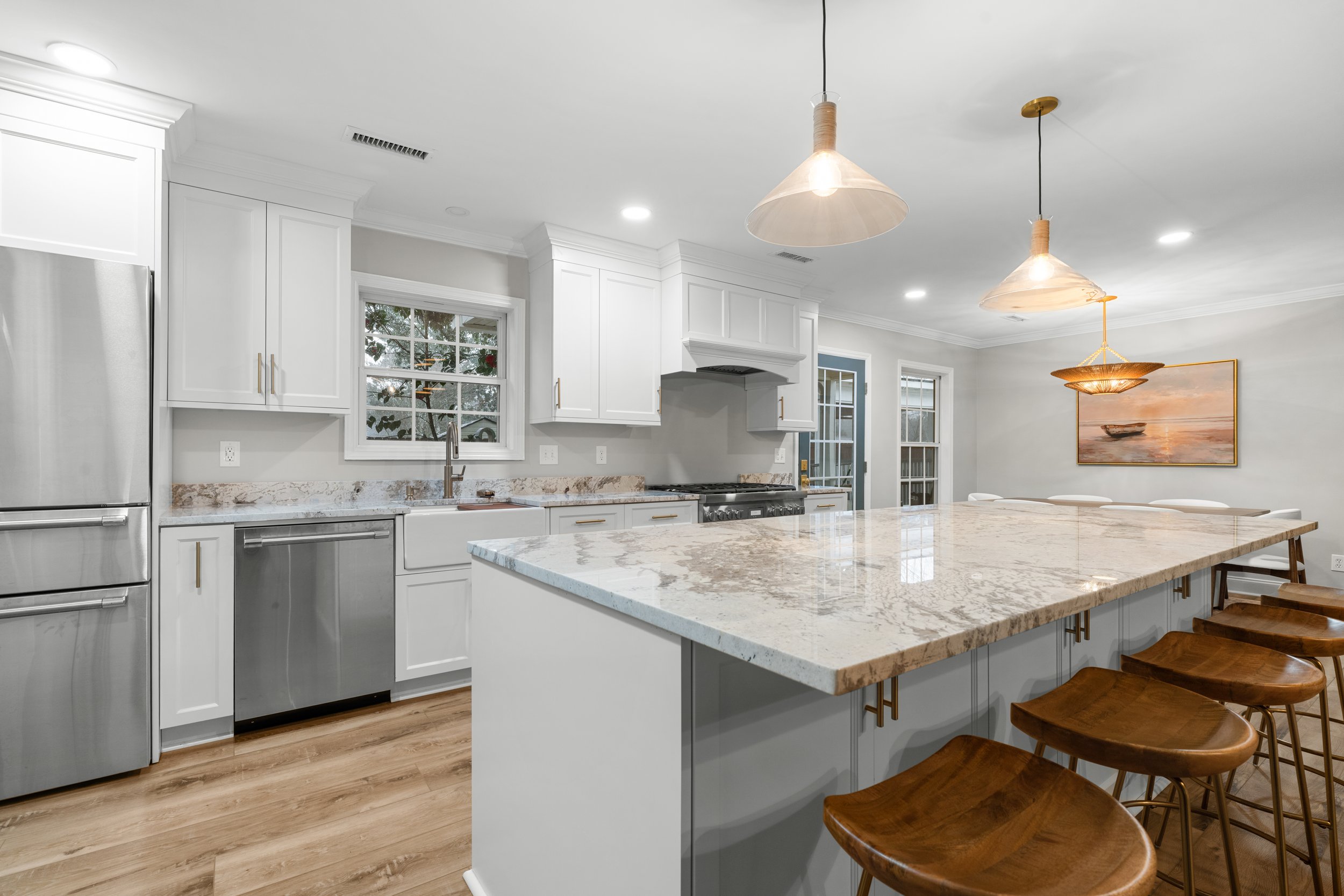 Modern kitchen with white cabinets, marble countertops, wooden bar stools, stainless steel appliances, pendant lighting, a painting on the wall, and hardwood flooring.
