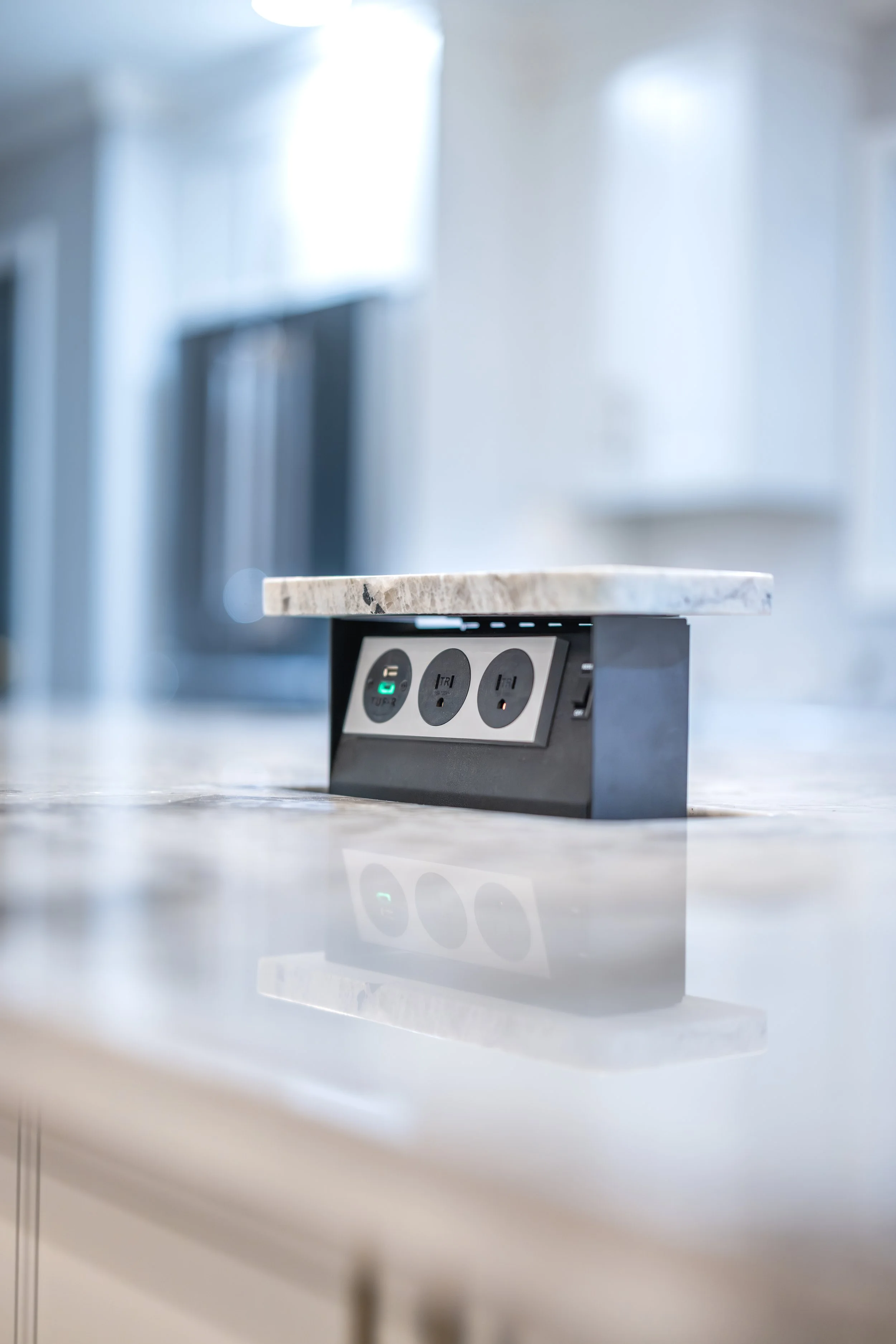 Kitchen countertop with a built-in power outlet and USB charging ports beneath a marble surface with a blurred background.