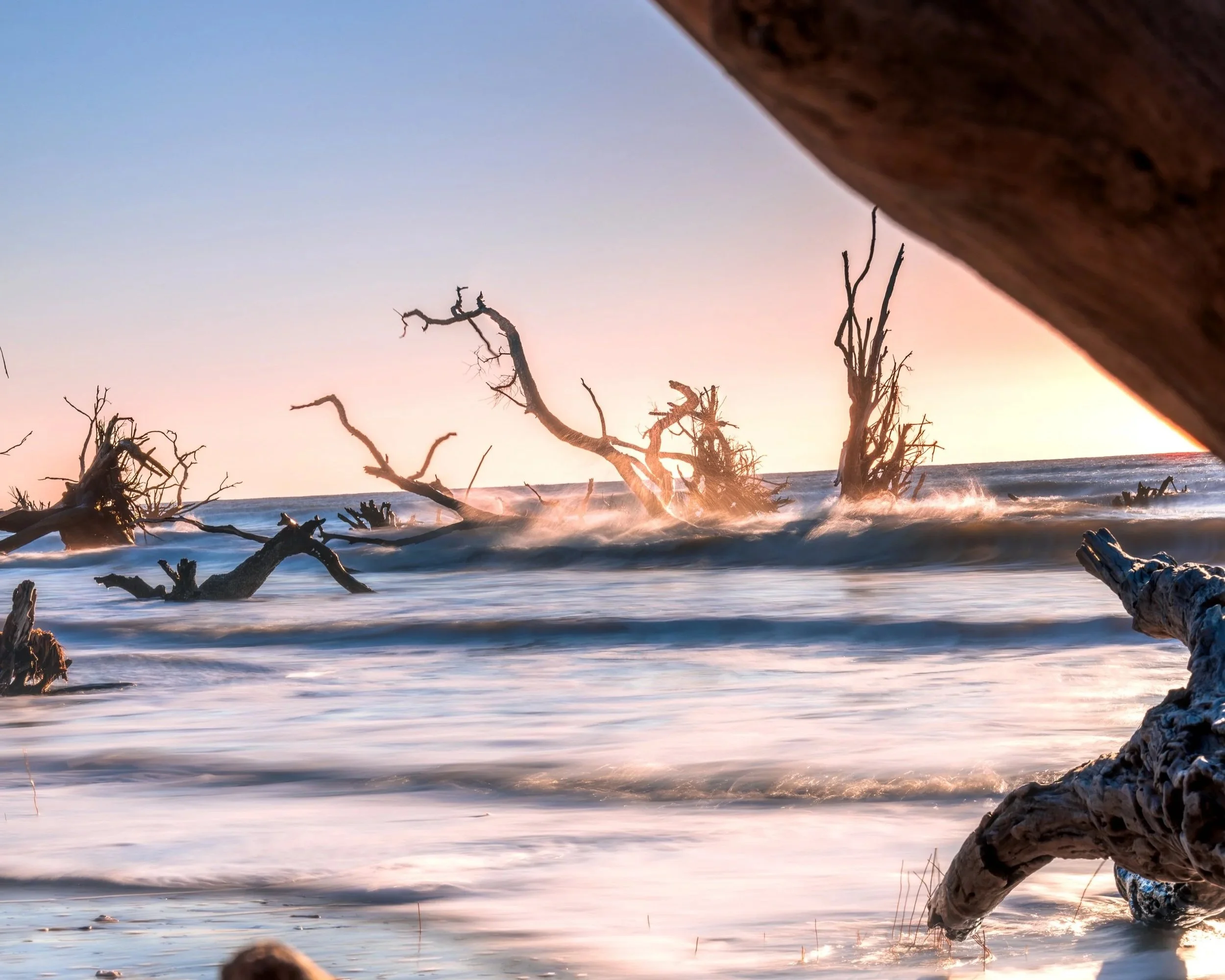 A sunset over the ocean with large, weathered driftwood on the shore and in the water, with some pieces partially submerged and others leaning against each other.