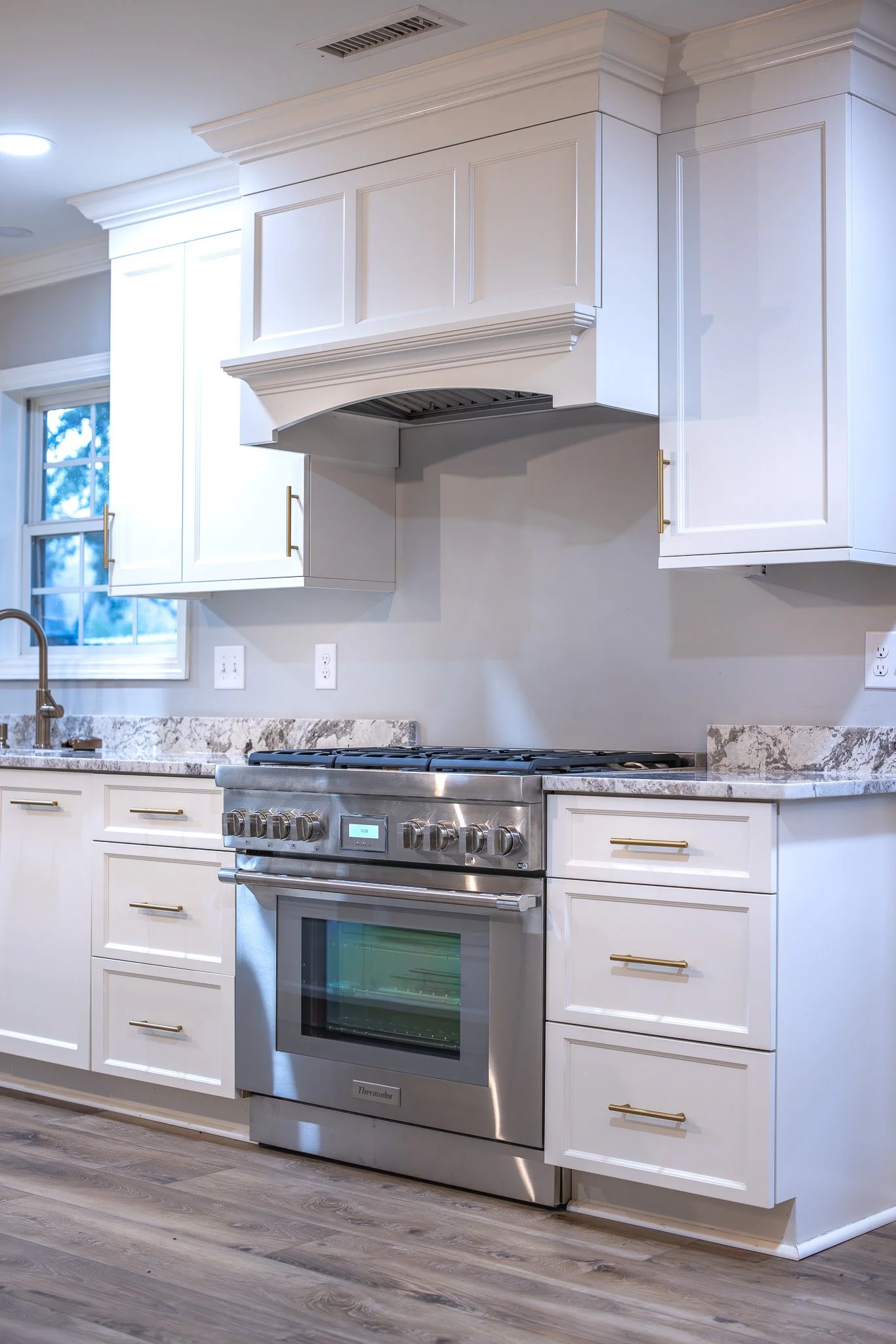 White kitchen with marble countertops, stainless steel oven, white cabinetry with gold handles, nearby window, and wooden floor.