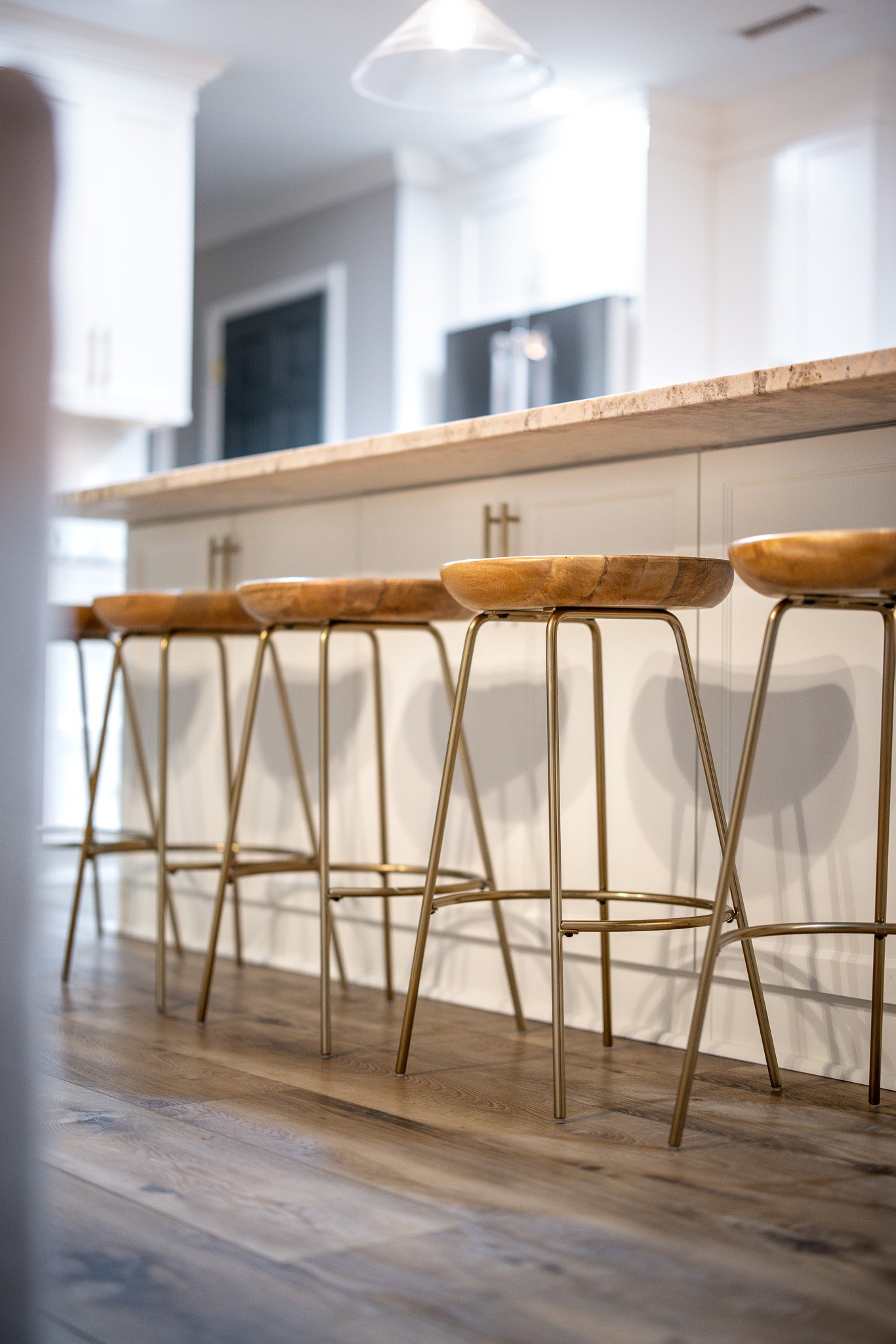 Close-up of a row of four modern bar stools with wooden seats and gold-colored metal legs in a bright, contemporary kitchen with white cabinetry and a marble countertop.
