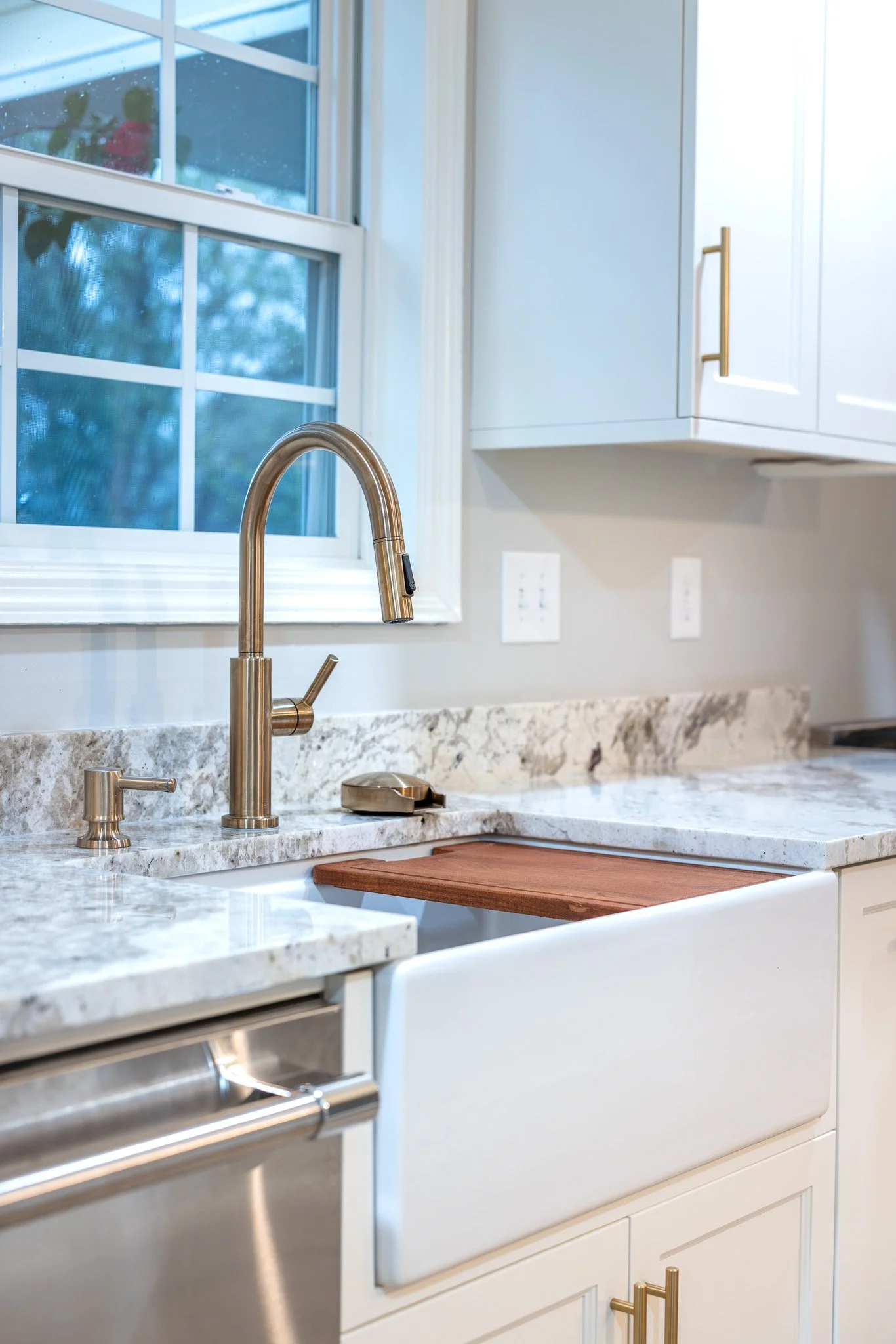 A modern kitchen with a marble countertop, a brass faucet, a window, and white cabinets with brass handles.