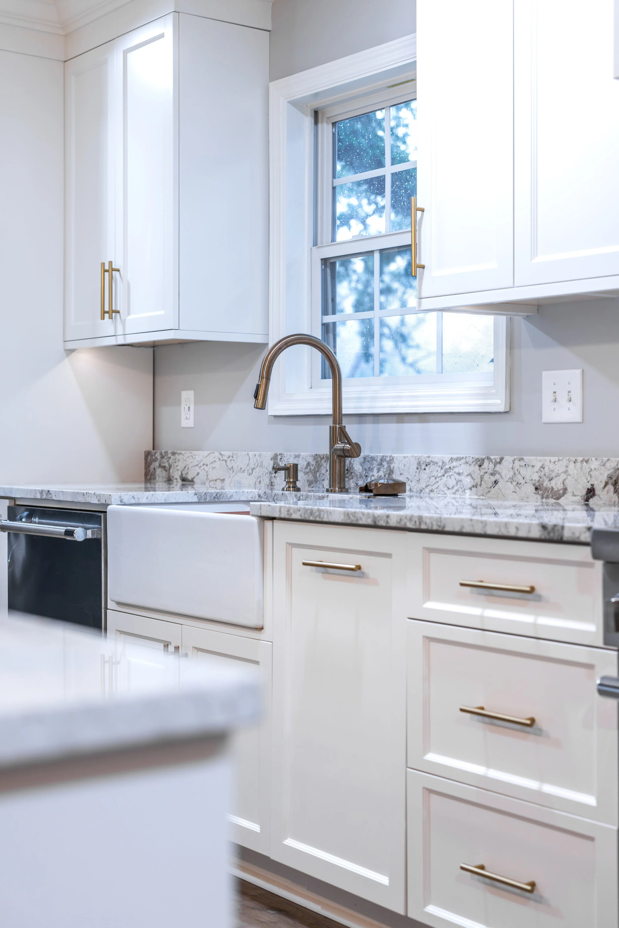 Kitchen with white cabinets, granite countertop, farmhouse sink, brass handles, window showing a rainy view outside.