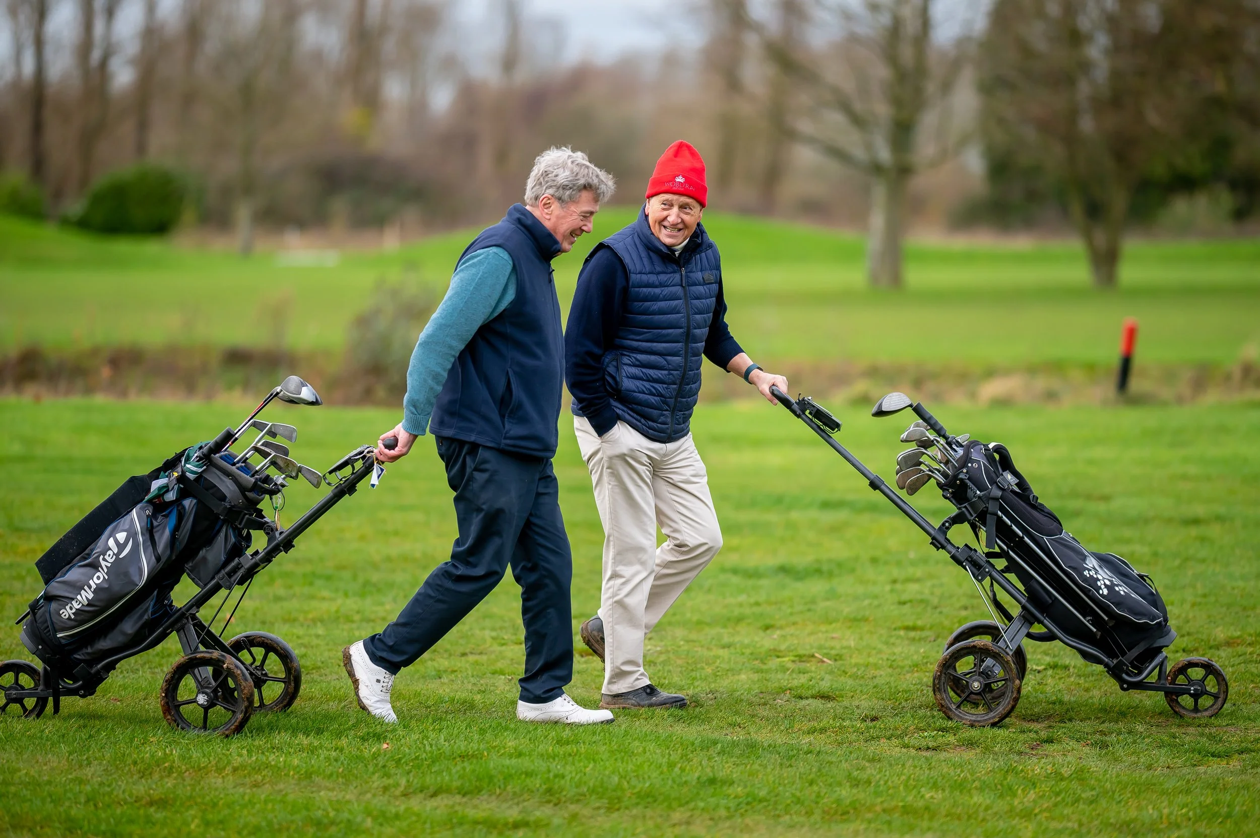 Two older men walking on a golf course, each pushing a golf cart with clubs, smiling and engaging in conversation.