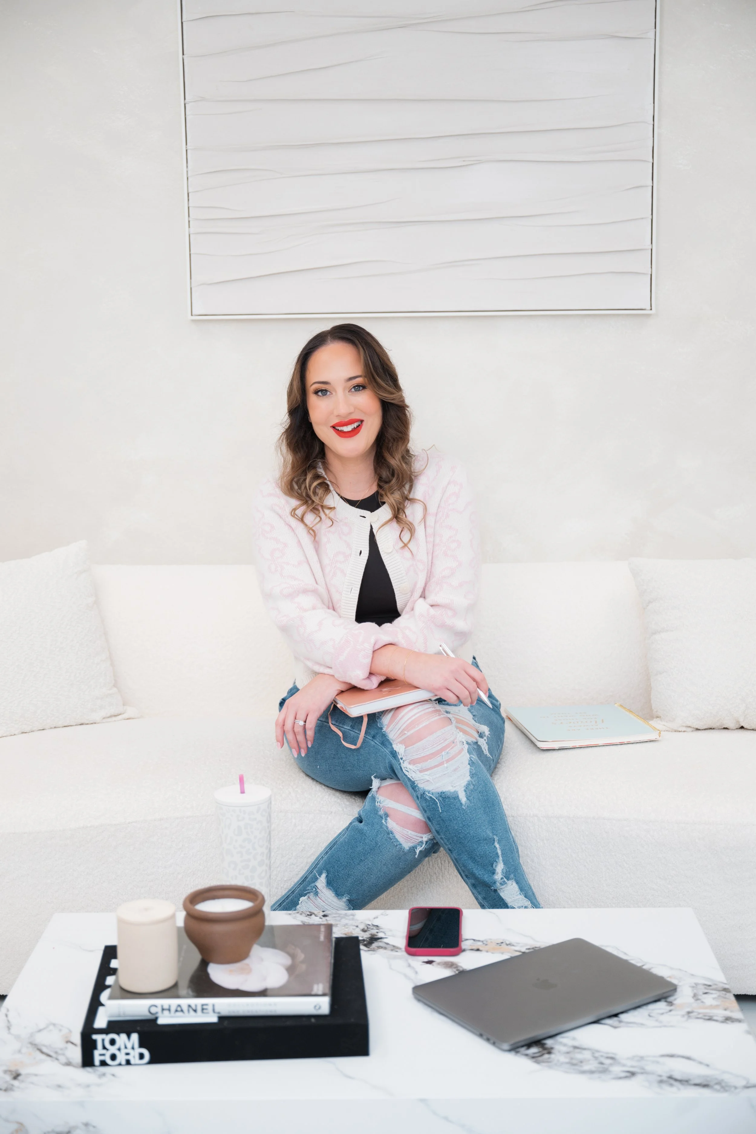 Attorney Tiffany Ann Jones sitting on a white sofa with a notepad and laptop on a marble coffee table, wearing a pink sweater and ripped jeans, against a light wall with abstract art.