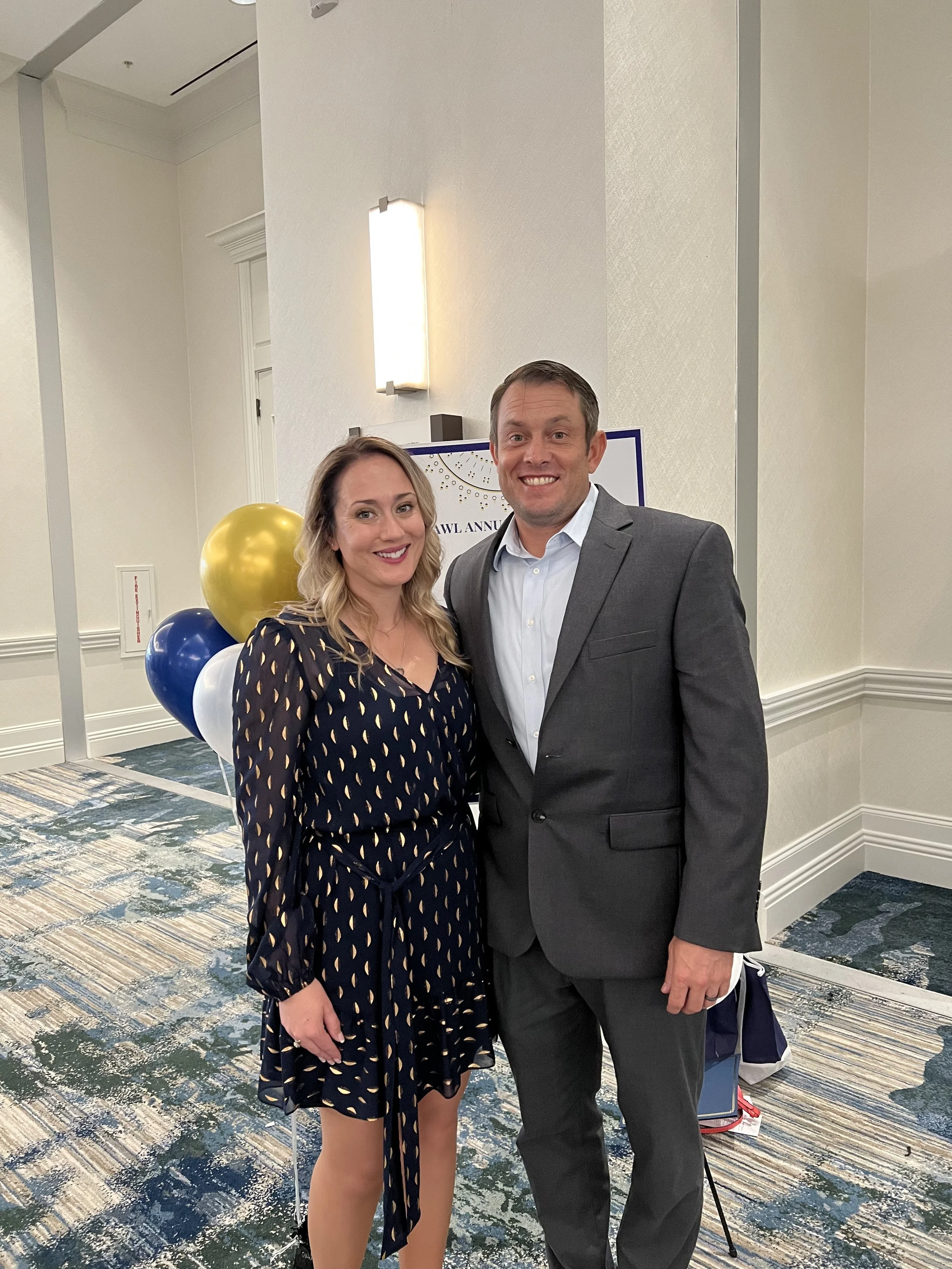 Tiffany Ann Jones and her husband posing together at the 2022 Florida Association for Women Law event. Tiffany is wearing a dark patterned dress and her husband is in a gray suit. They are standing on a carpeted floor with balloons in the background.