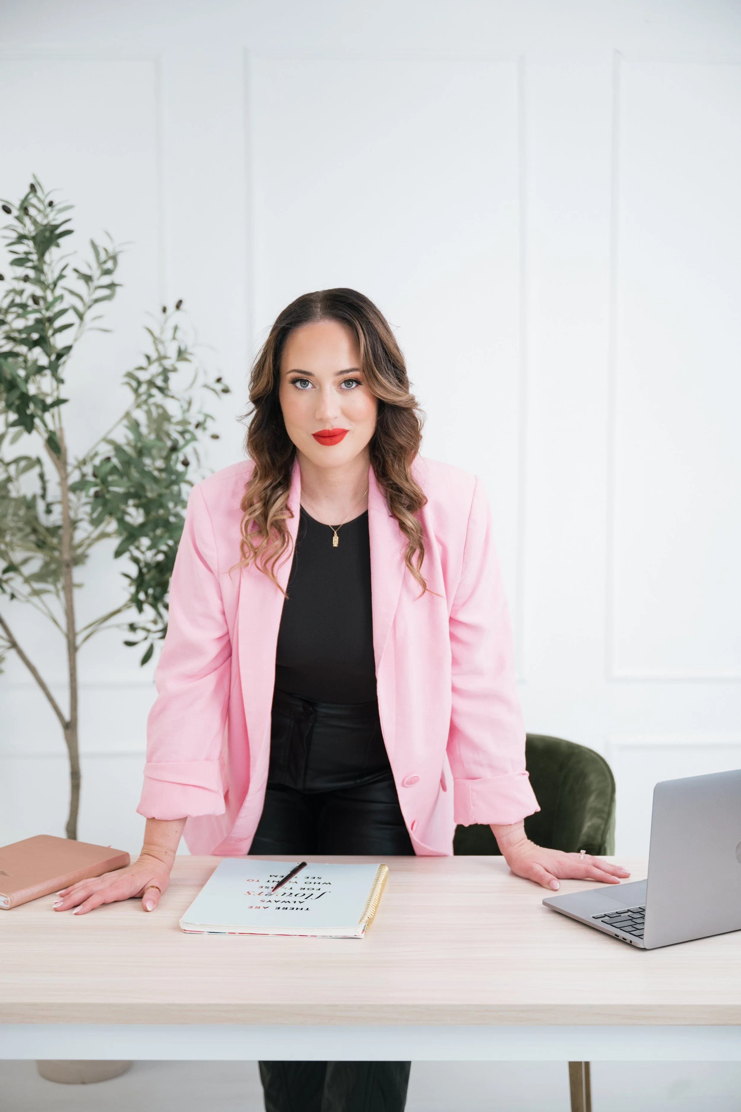 Attorney Tiffany Ann Jones in a pink blazer standing at a desk with a laptop, notebook, and plant in the background.