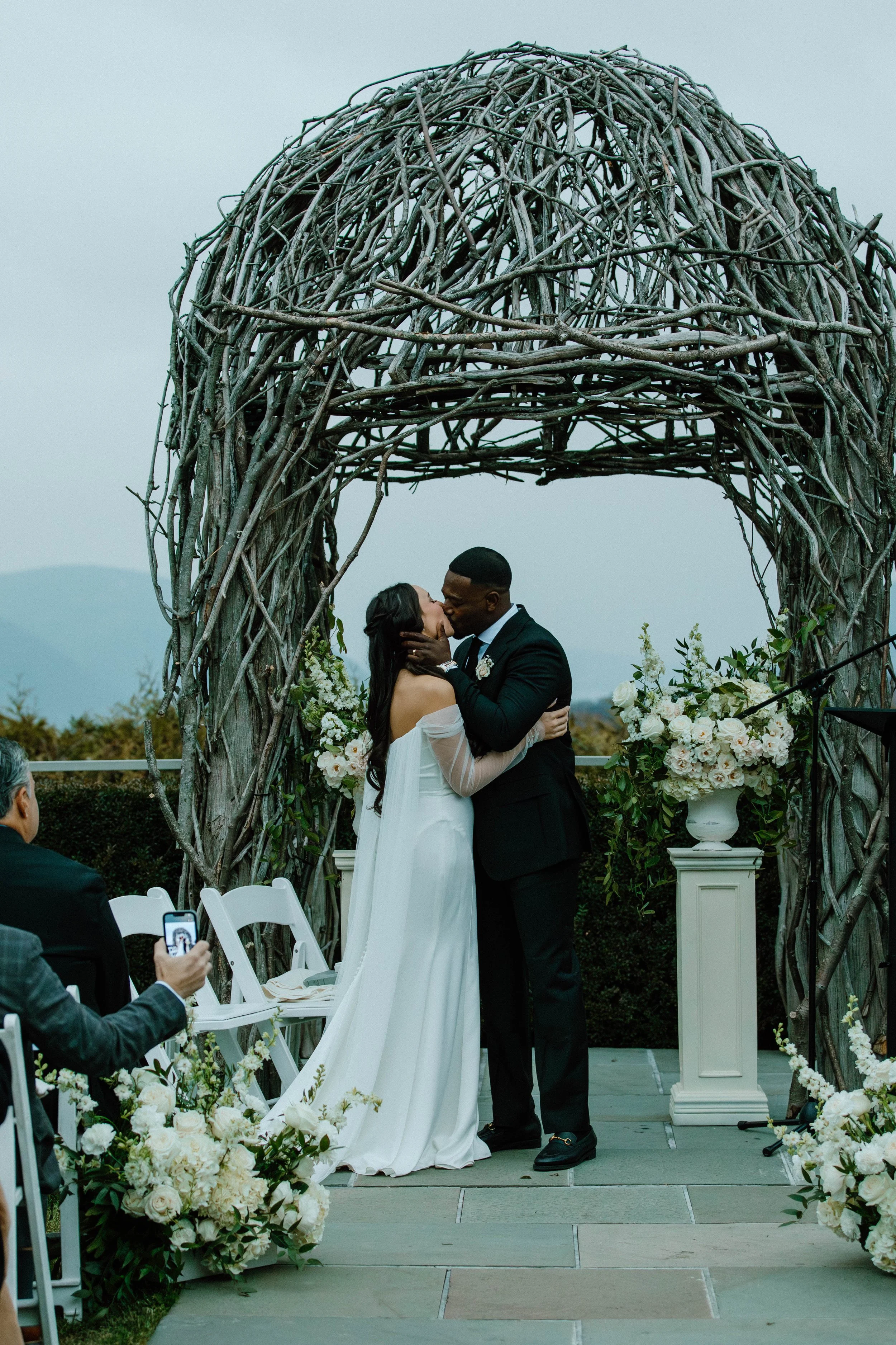 Bride and groom kissing under the ceremony pergola at The Garrison in the Hudson Valley with the Hudson Highlands in the background and beautiful white floral arrangements on pedestal stands next to them
