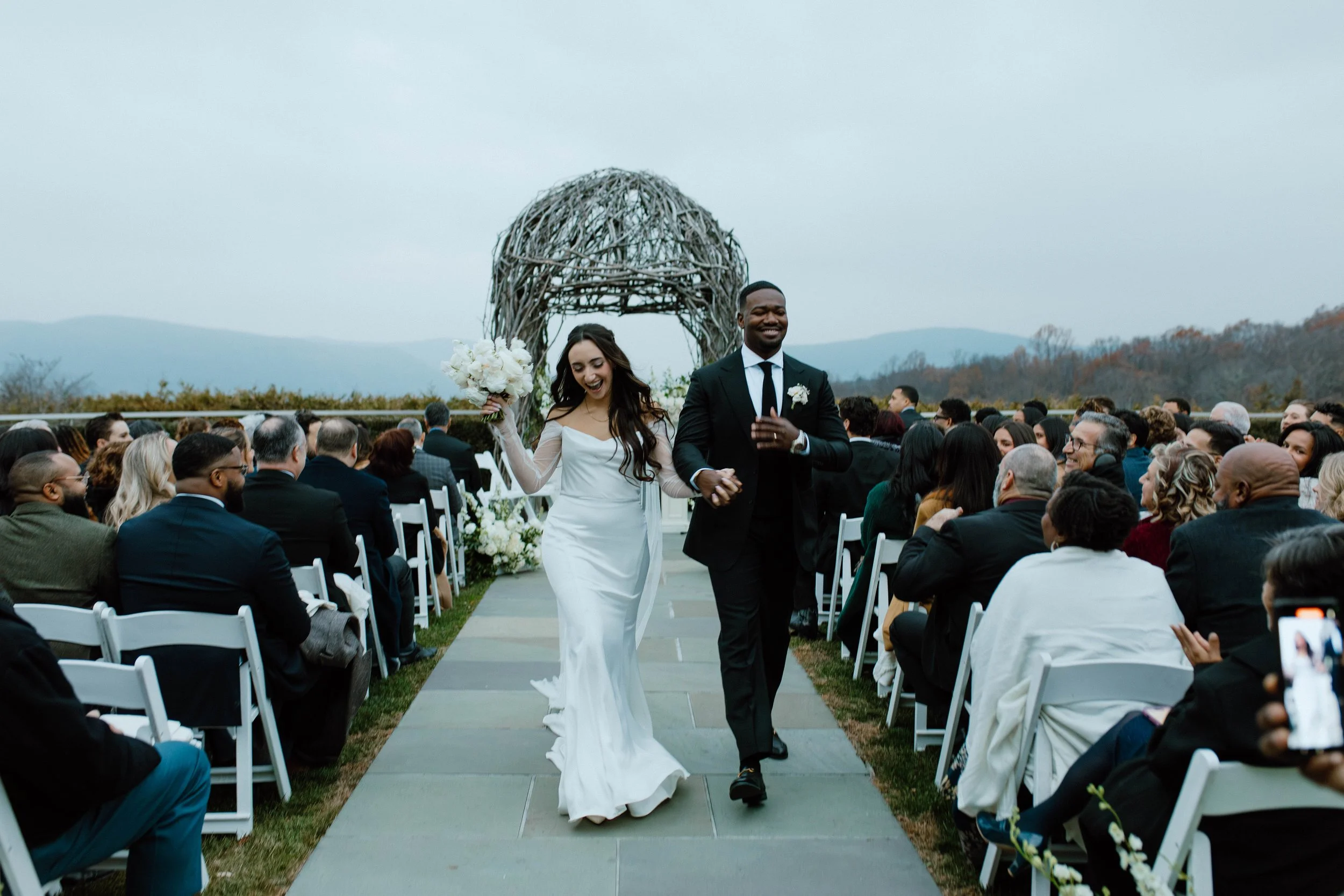 Bride and groom walking down the aisle holding bouquet at their ceremony at The Garrison in the Hudson Valley with mountains as the background in fall