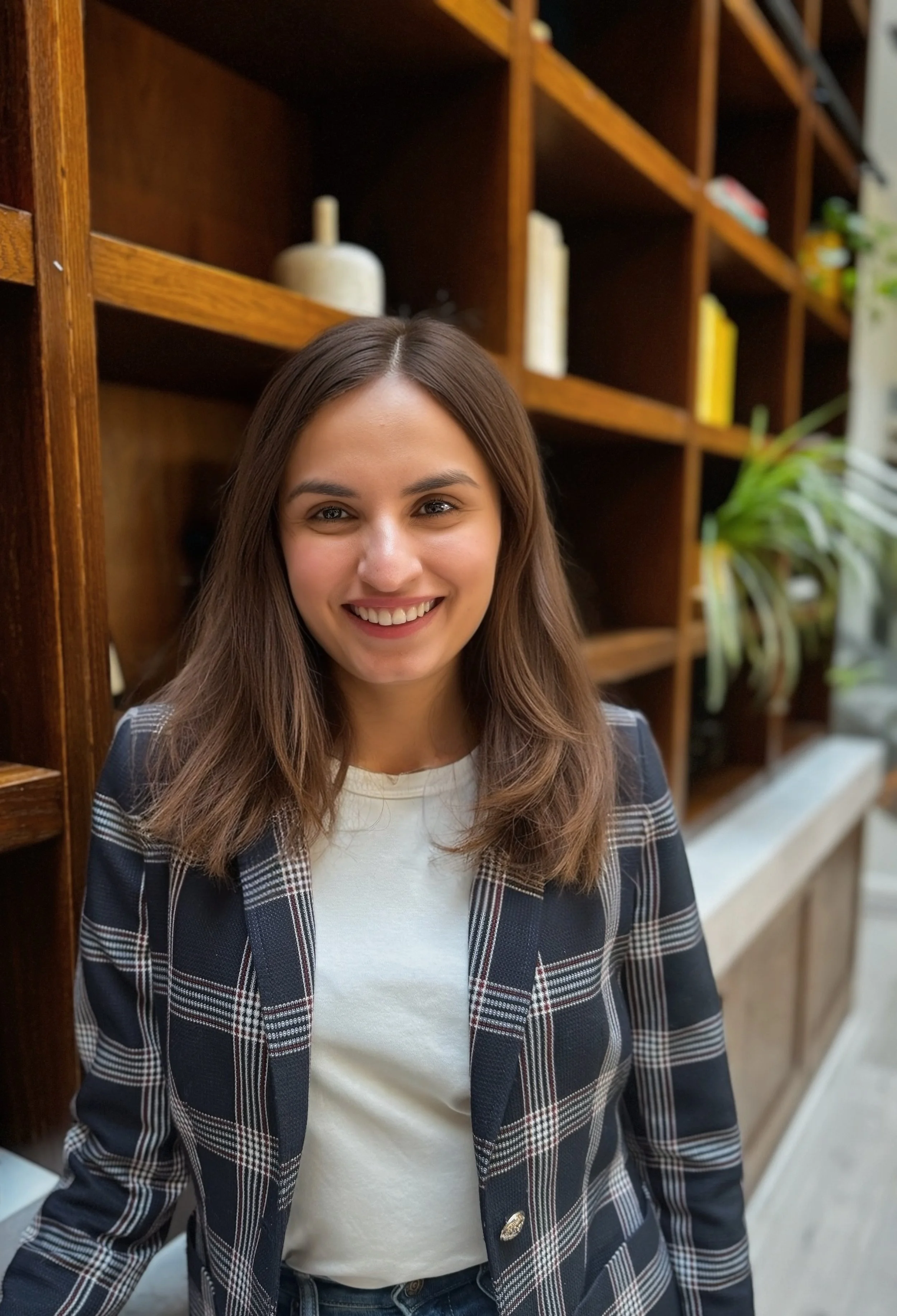 A woman with long brown hair, wearing a white shirt and plaid blazer, standing in front of a wooden bookshelf with books and decorative objects.