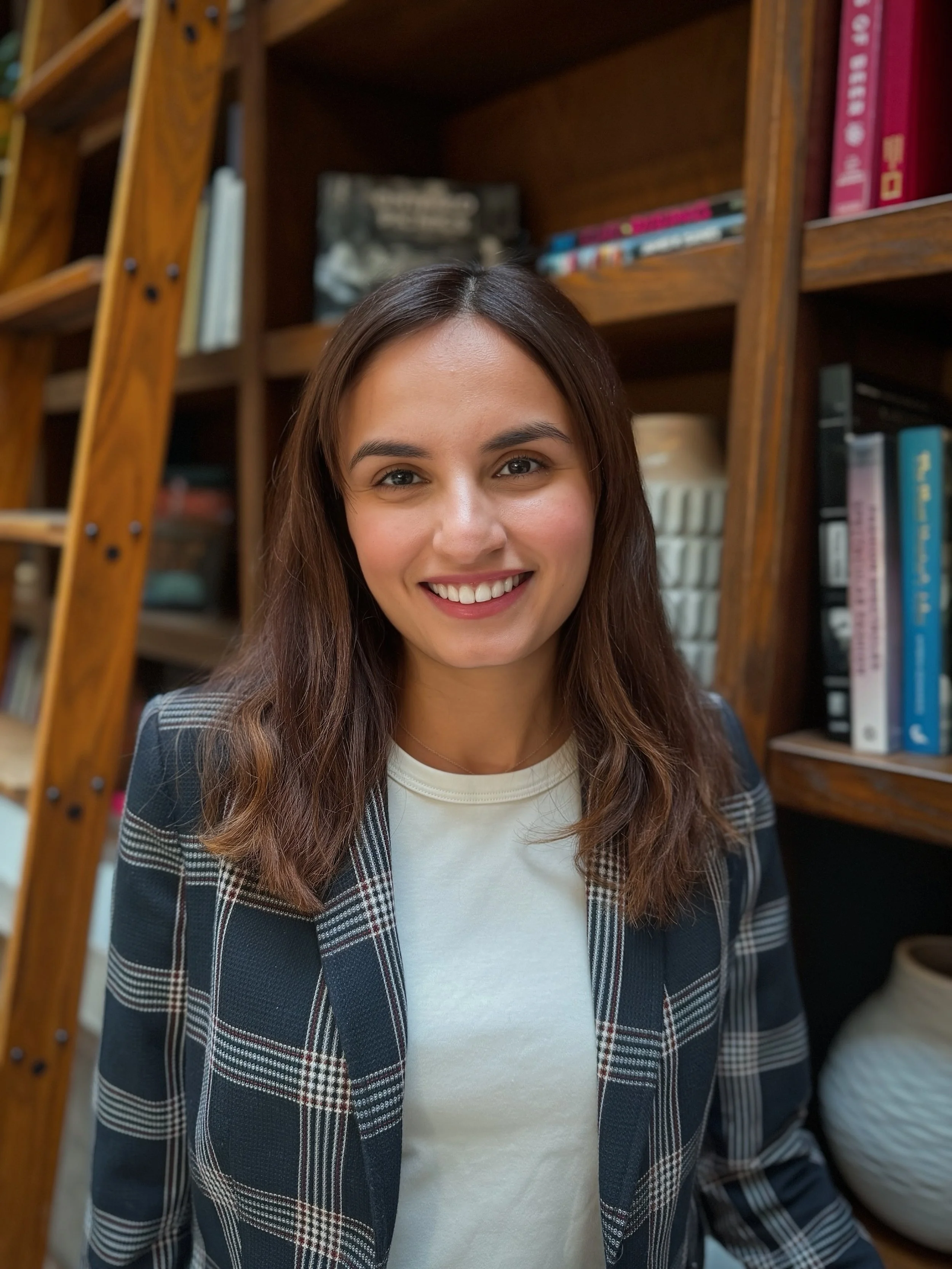 Young woman with long brown hair smiling, standing in front of a wooden bookshelf filled with books.