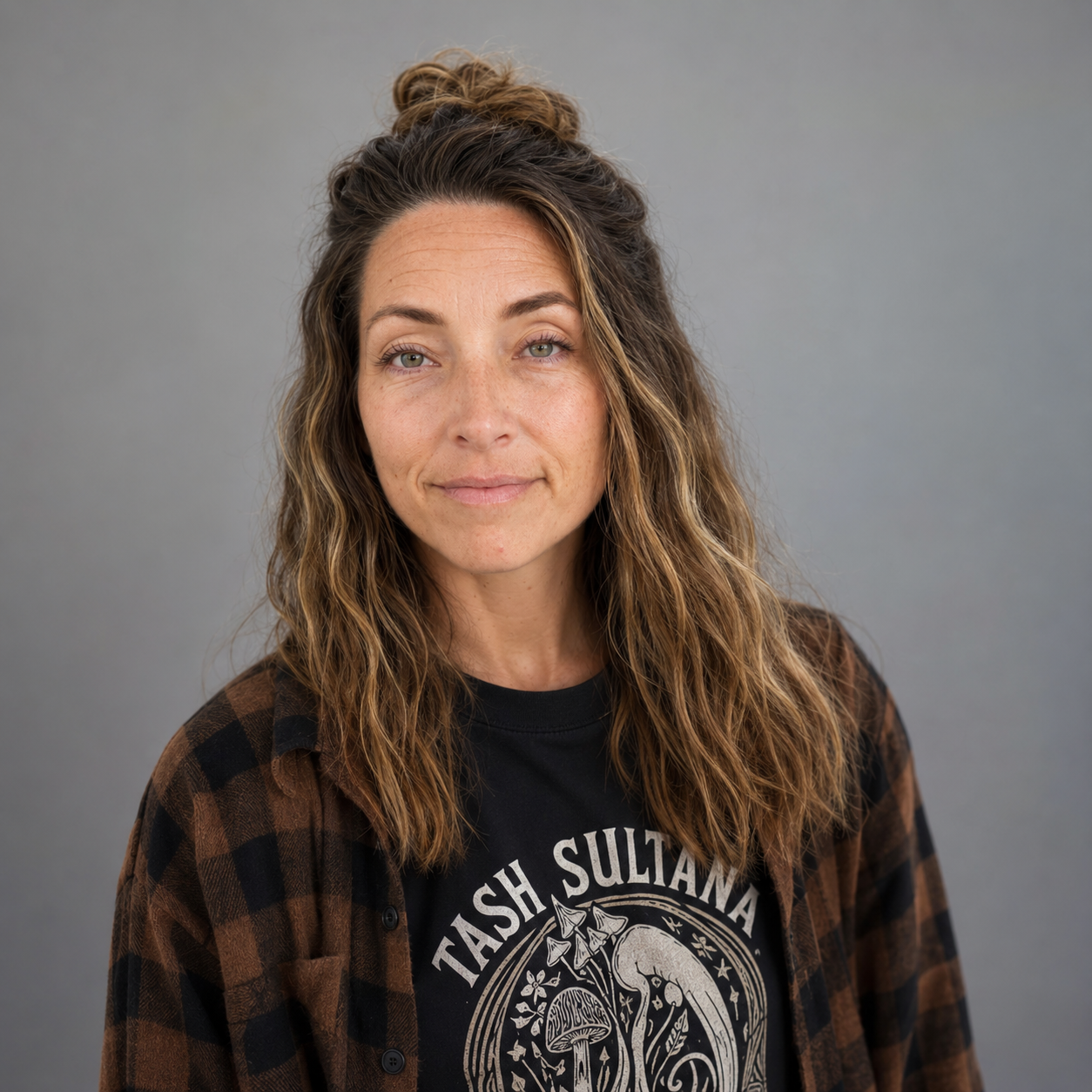 A woman with long, wavy brown hair styled in a half-up bun, wearing a black T-shirt with a mushroom graphic and a brown checkered shirt, standing against a plain grey background.