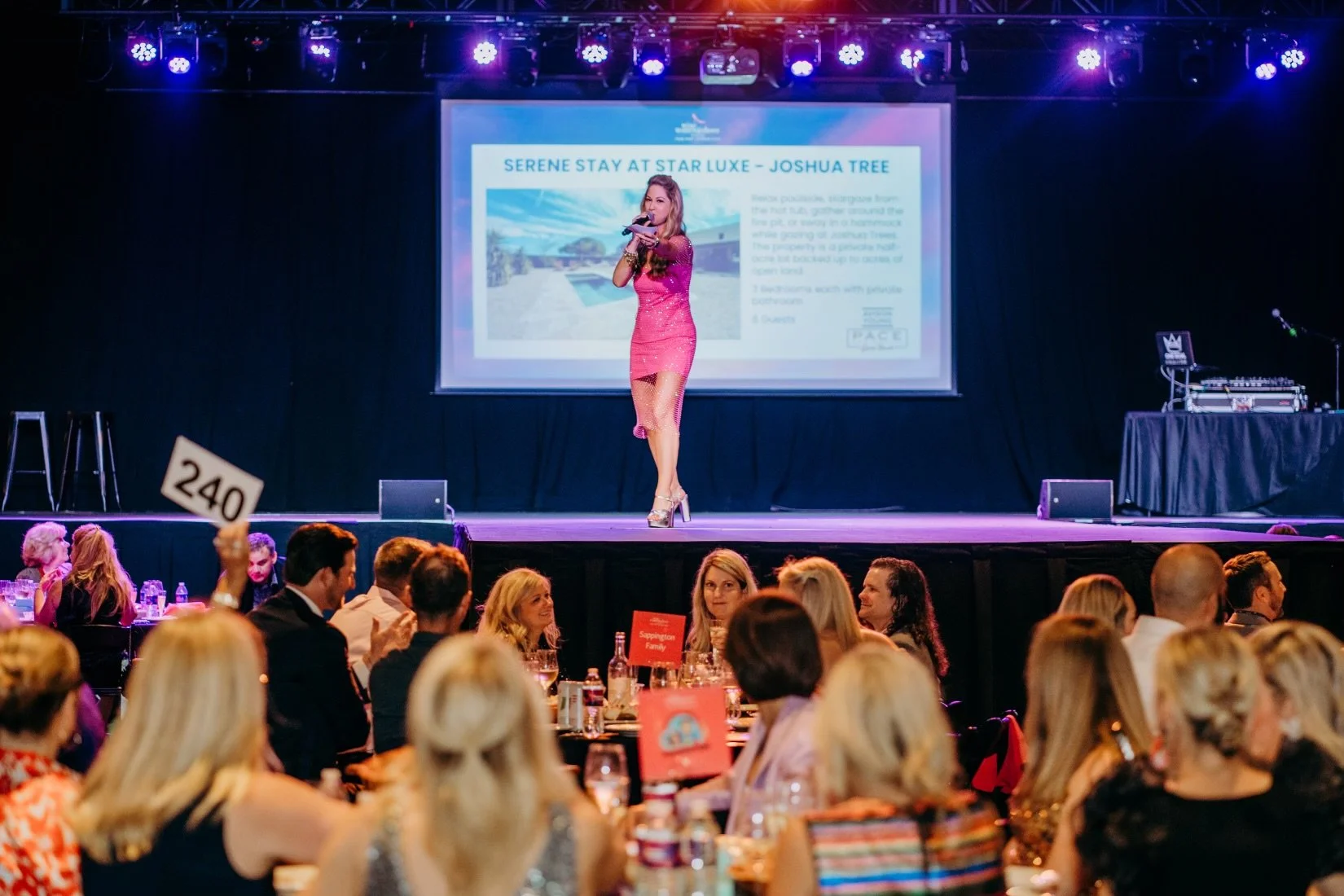 Ashlee Meier - Champion of Joy. A woman in a pink dress performs on stage at a formal event, with an audience seated at round tables watching her. The stage has a large screen behind her displaying a slide with text and an image.
