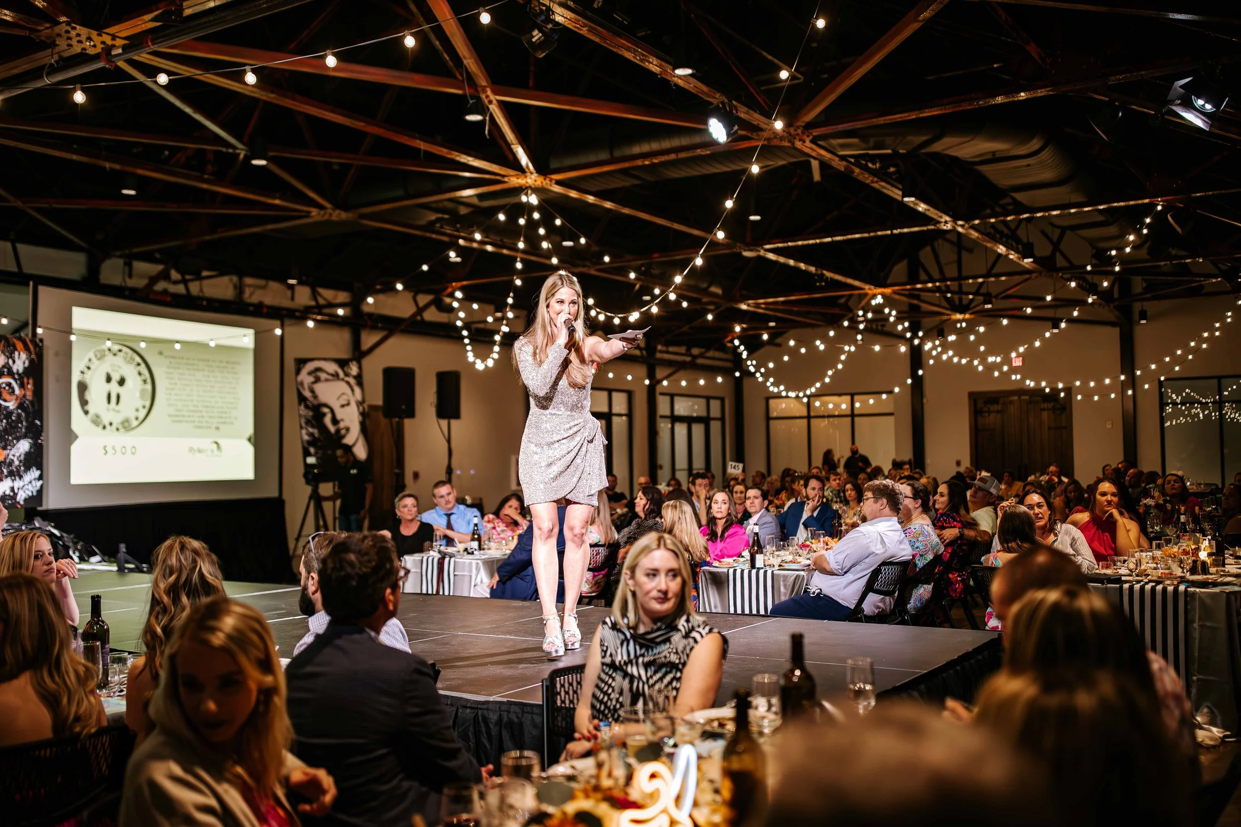 A woman in a silver dress is on stage speaking into a microphone at a large indoor event with many seated guests, string lights hanging from the ceiling, and a projector screen in the background.