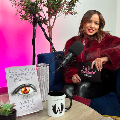 Young woman sitting on a blue couch with a pink and white wall behind her, holding a podcast sign. In front of her are a book titled 'A Journey to Becoming the Best-Self,' a black microphone, a white mug with a black dragon logo, and a plant.