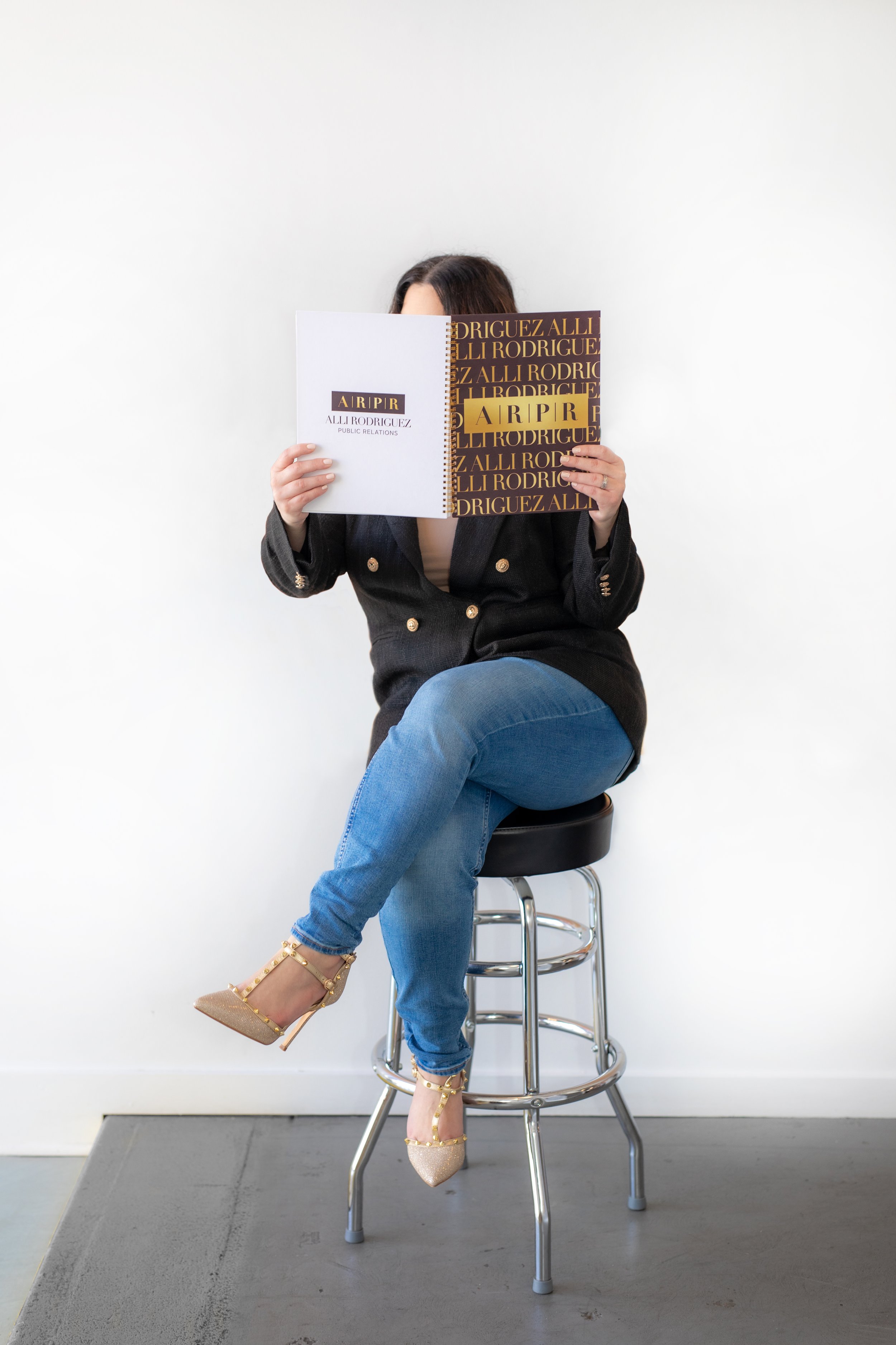 Person sitting on a stool, holding a magazine open in front of their face, wearing a black jacket, jeans, and studded high-heeled shoes.