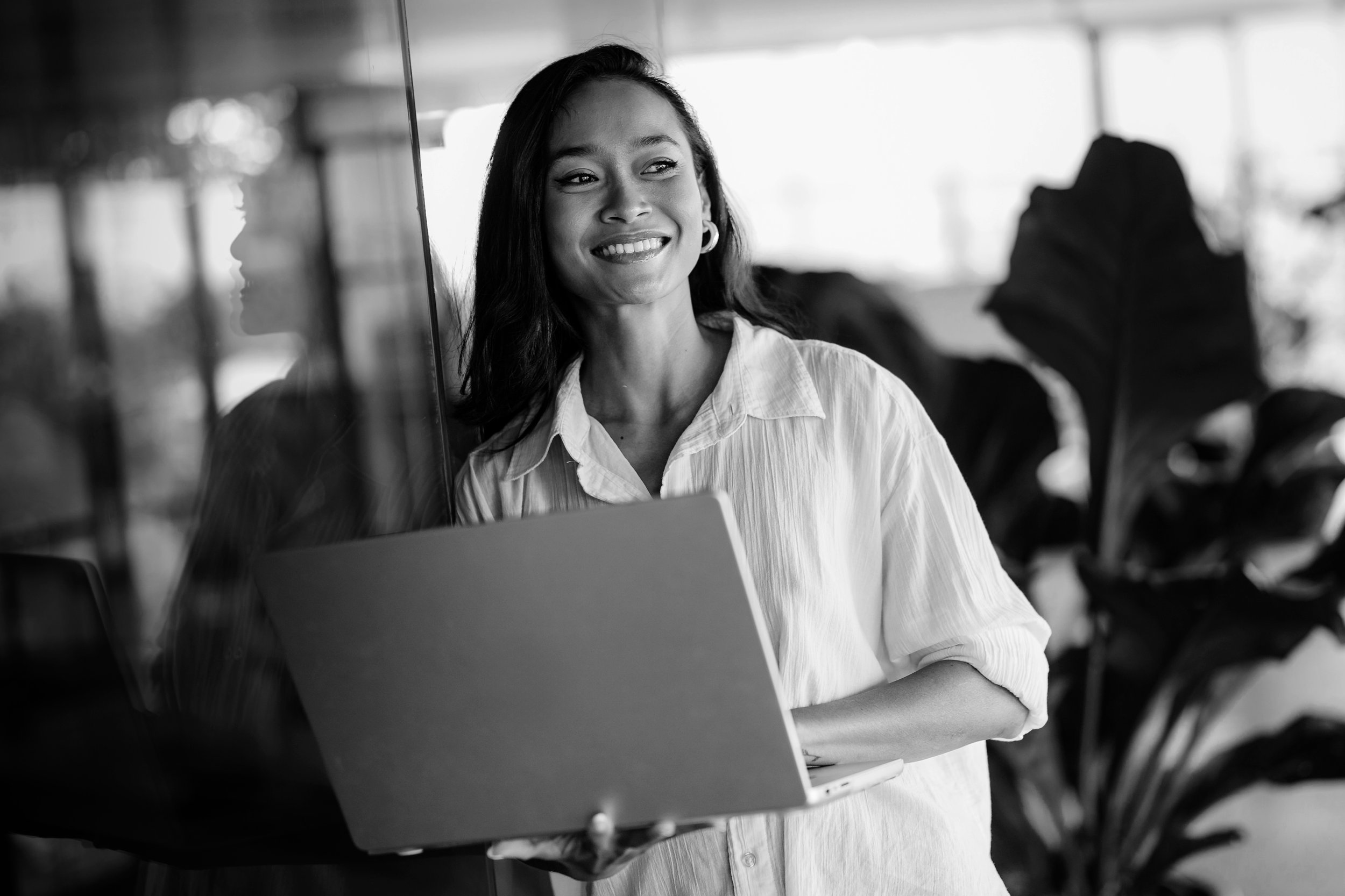 Mujer sonriendo con computadora portátil en mano, en un entorno interior con plantas, vista en blanco y negro.