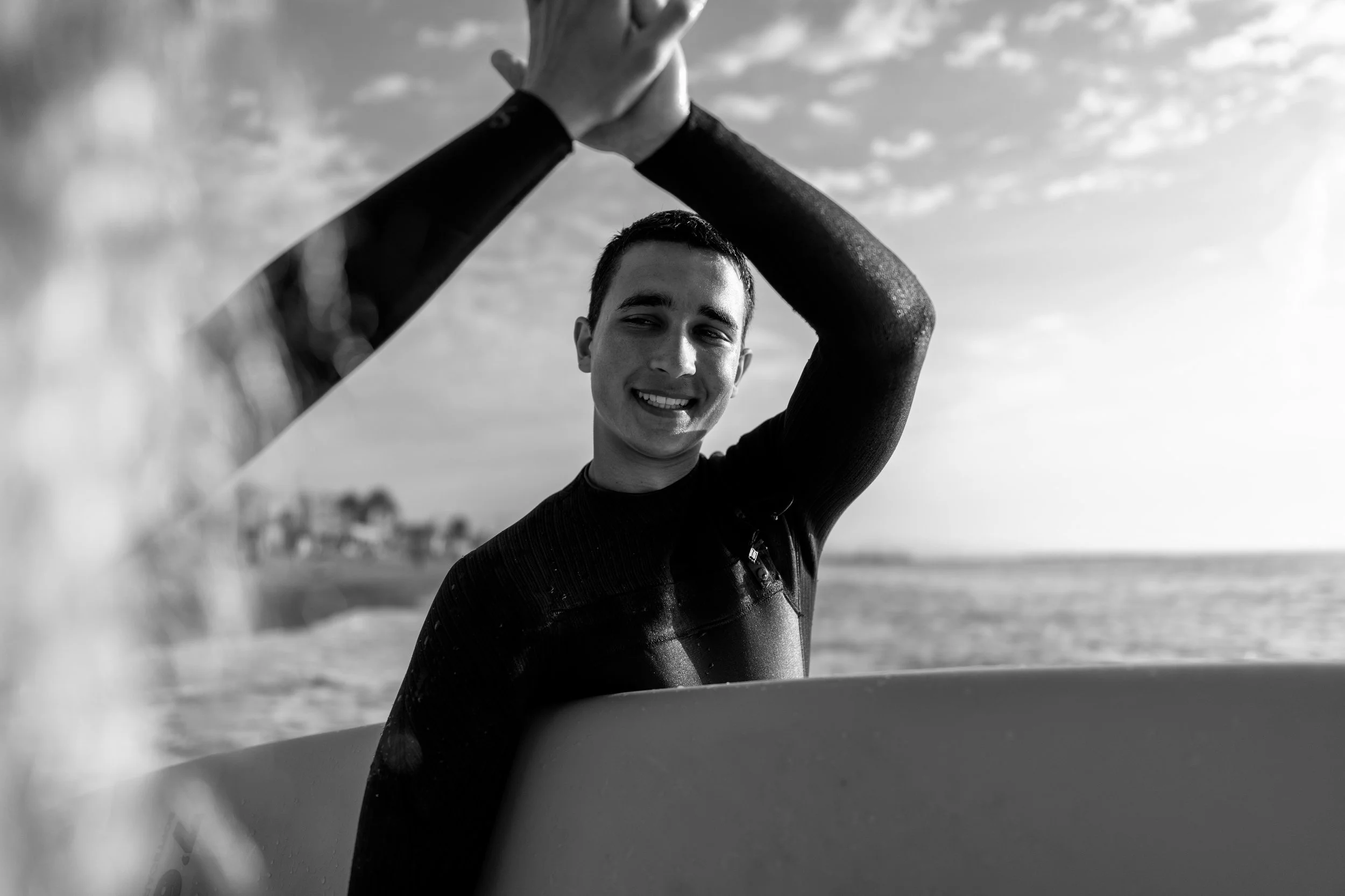 Persona sonriendo en la playa con traje de neopreno, levantando las manos, en negro y blanco, con el mar y cielo en el fondo.
